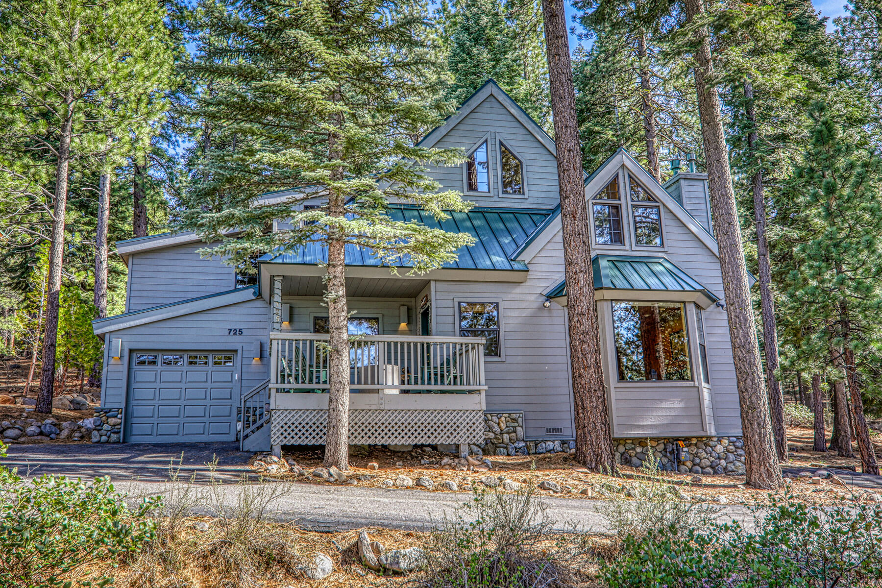 This charming, multi-level mountain home features light grey horizontal siding, a striking green metal roof, and stone foundation accents. A welcoming wooden deck and a built-in garage add to the property's curb appeal, while tall, mature pine trees provide a serene and authentic woodland setting.