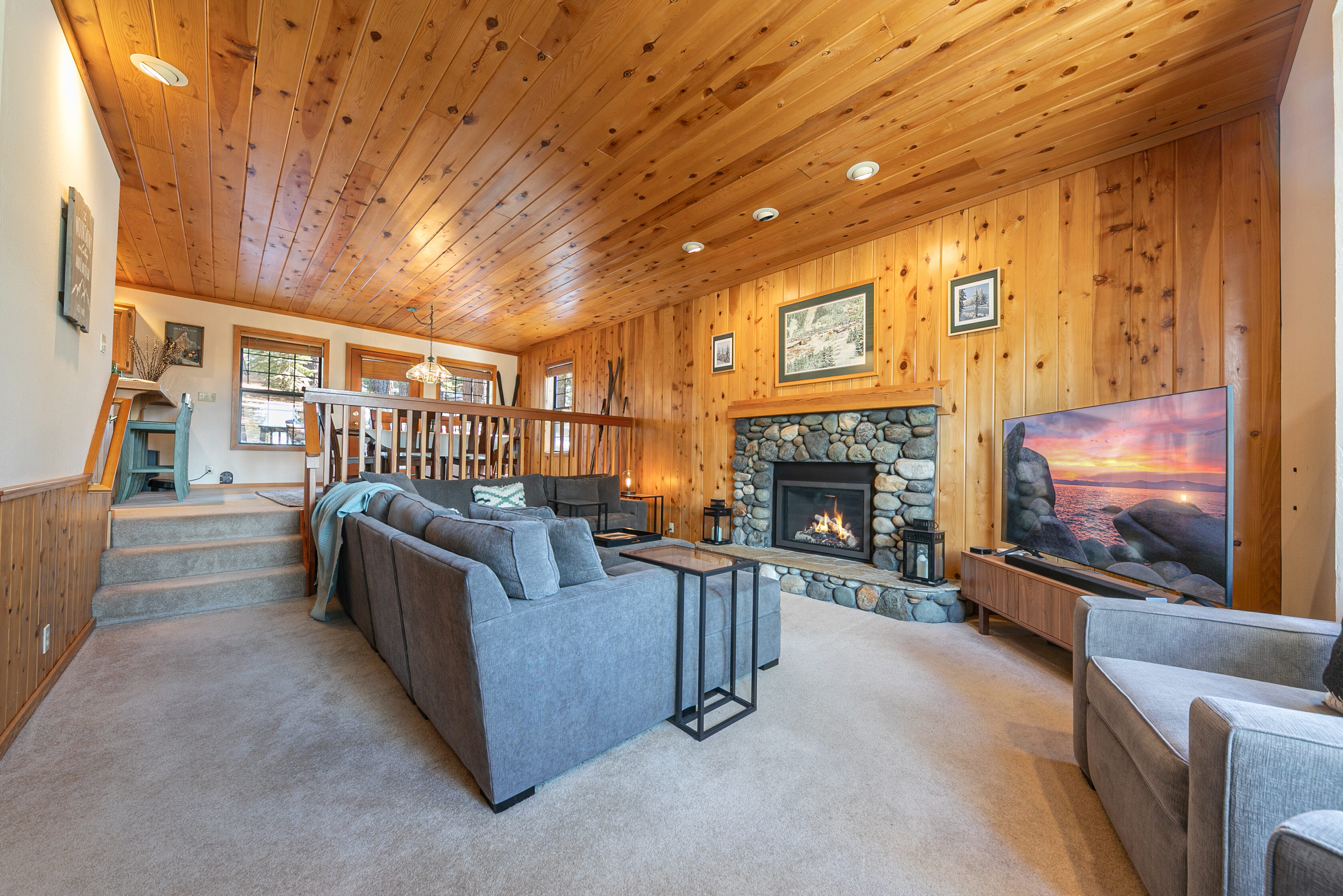 This cozy, mountain-style living room features warm wood-paneled walls and a stunning stone fireplace as the central focal point. The space is furnished with a large, comfortable gray sectional sofa and a sleek media console, creating a welcoming atmosphere for relaxation. With an open-concept layout leading to a slightly elevated dining area, the room offers both functionality and classic cabin charm.