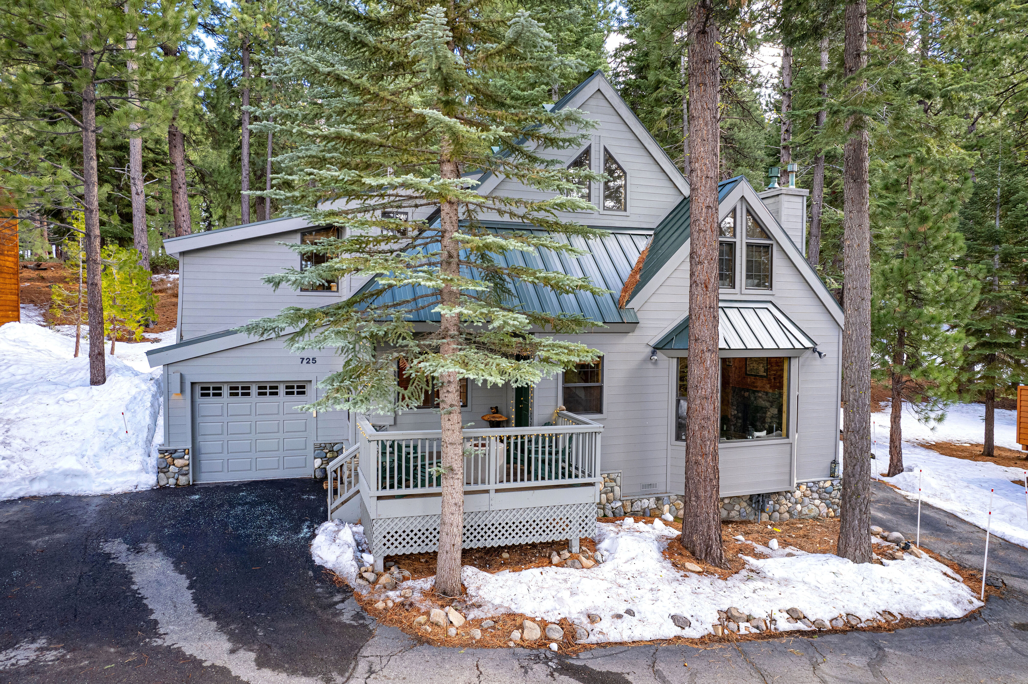 This charming, multi-level mountain cabin features gray siding, a striking dark green metal roof, and stone foundation accents, set within a serene, snow-dusted pine forest. The front of the home offers a welcoming raised wooden deck and a two-car garage, while the architectural design includes multiple gables and large windows that fill the home with natural light. Its rustic, cozy aesthetic is perfectly suited for a peaceful mountain getaway or permanent residence.