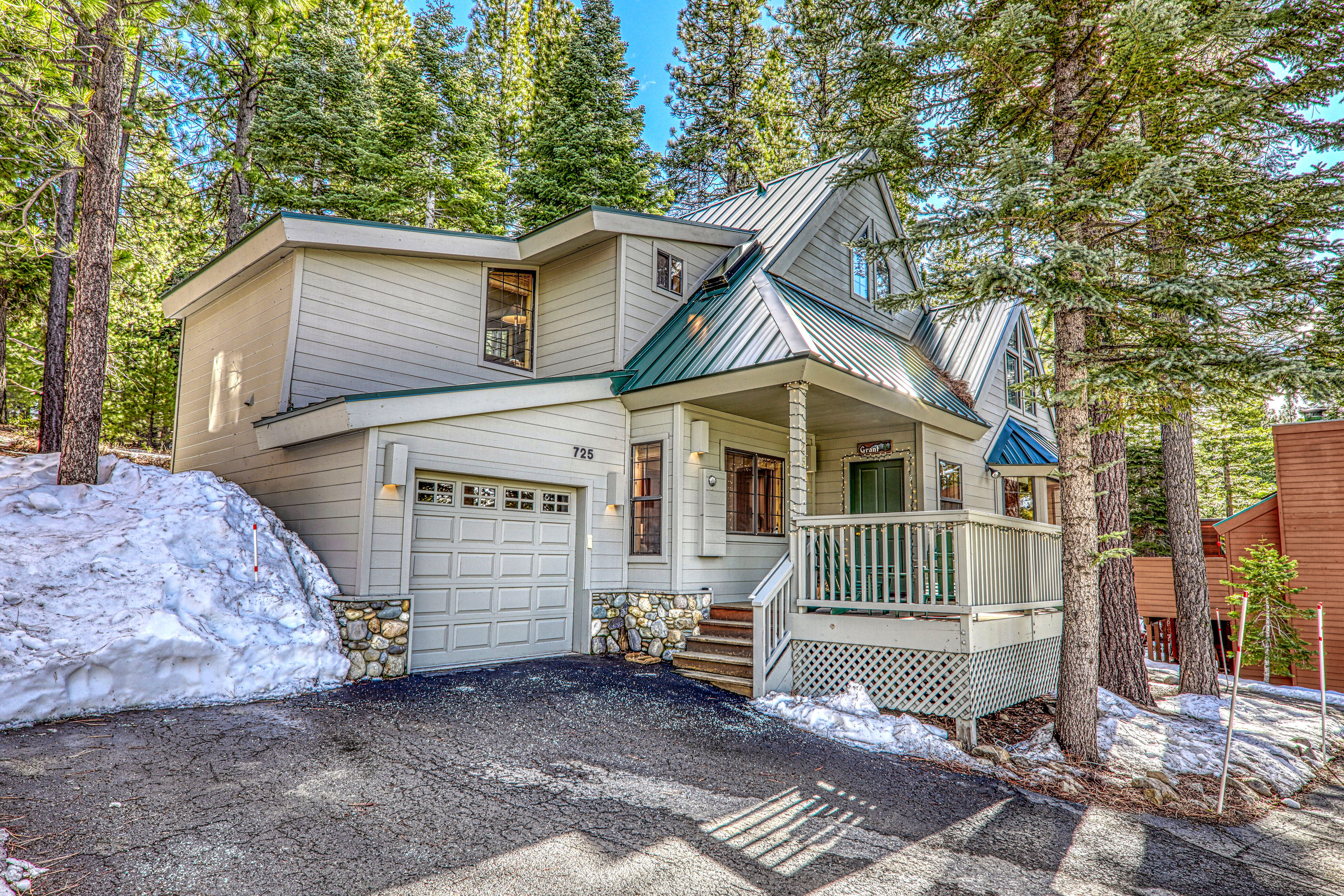 This is a charming mountain-style home featuring light gray horizontal siding, a striking green metal roof, and stone accents along the foundation. The property includes a paved driveway, an attached garage, and a welcoming covered porch, all set in a scenic, forested location with remnants of snow on the ground. Its rustic yet well-maintained aesthetic makes it a perfect retreat for mountain living.