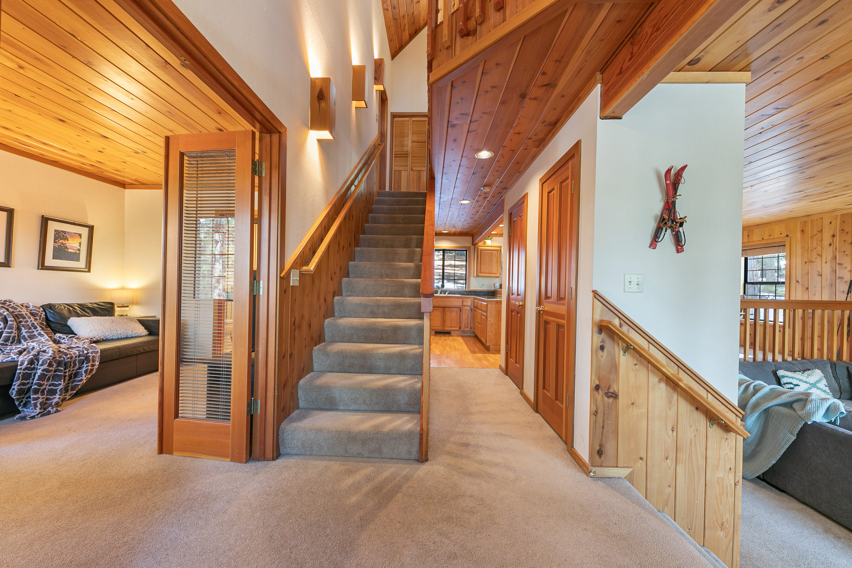 This cozy mountain-style home features a central carpeted staircase as the core of its floor plan, accented by warm wood paneling and ambient wall sconces overhead. The space flows seamlessly between a comfortable living room with a leather sofa, a functional kitchen visible in the background, and an additional lower living area characterized by rustic lodge decor, including decorative vintage skis on the wall. Its combination of natural wood accents and multi-level layout creates a warm, inviting, and well-organized atmosphere.