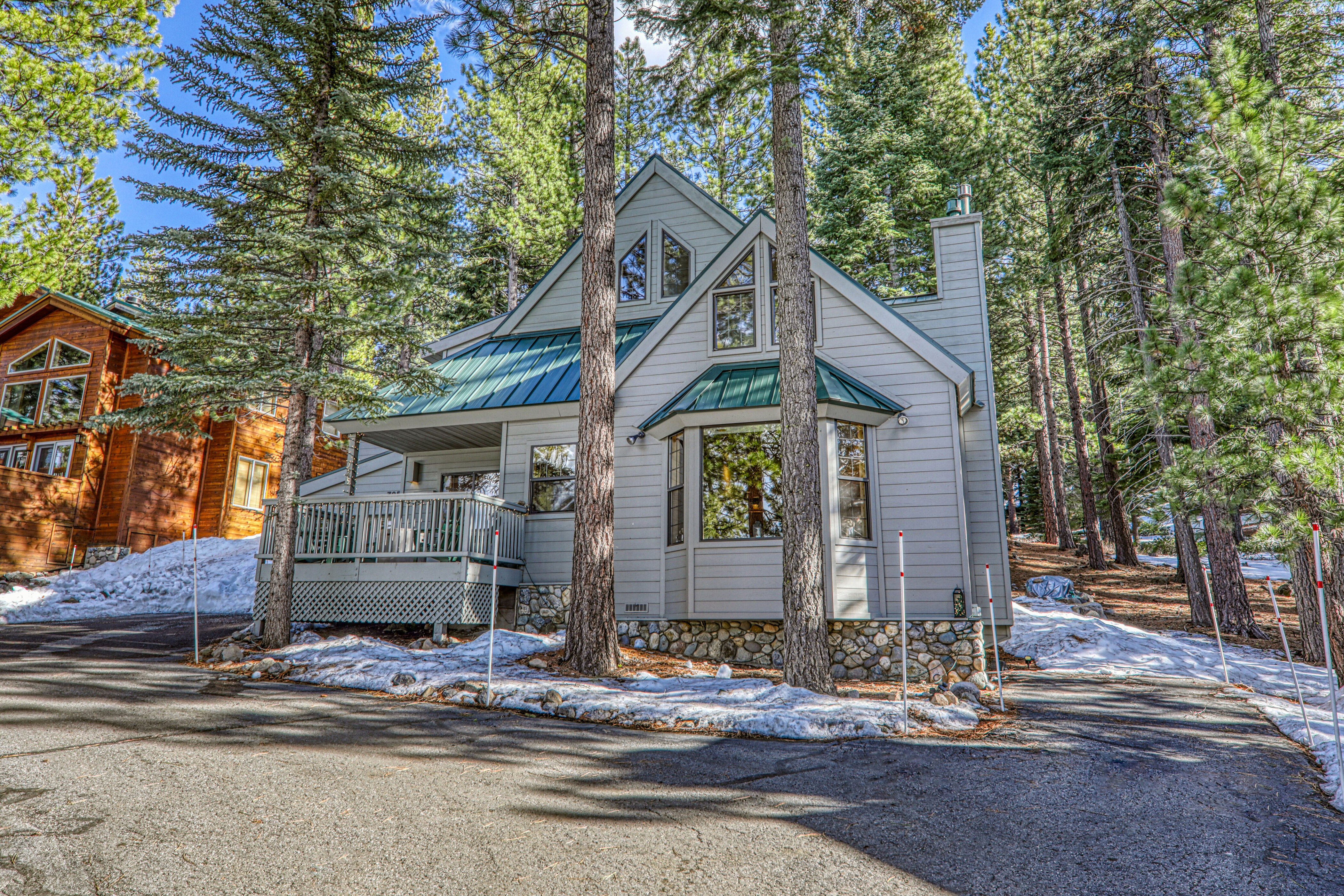 This charming, multi-level mountain cabin features light gray siding with a bold green metal roof and a stone foundation. The exterior includes a wooden deck, multiple windows including a prominent bay window, and a driveway lined with snow and tall pine trees. The home offers a classic, cozy aesthetic perfect for a wooded, alpine setting.