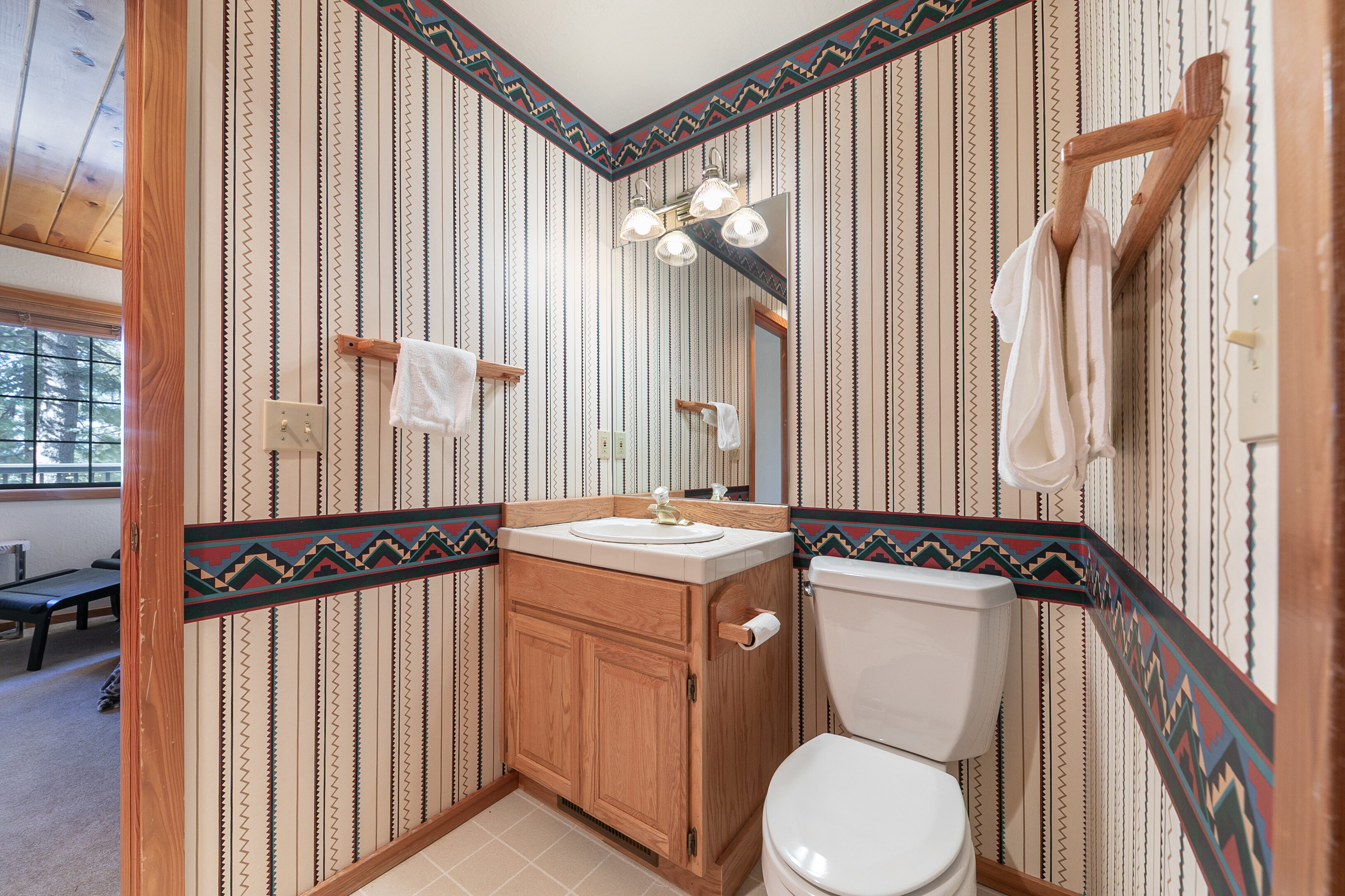 This compact half-bathroom features a distinct Southwestern-themed wallpaper with geometric patterns and vertical stripes. The space is efficiently designed with a light oak vanity, a neutral tiled countertop, and a classic white toilet, all complemented by warm wood trim and hardware.