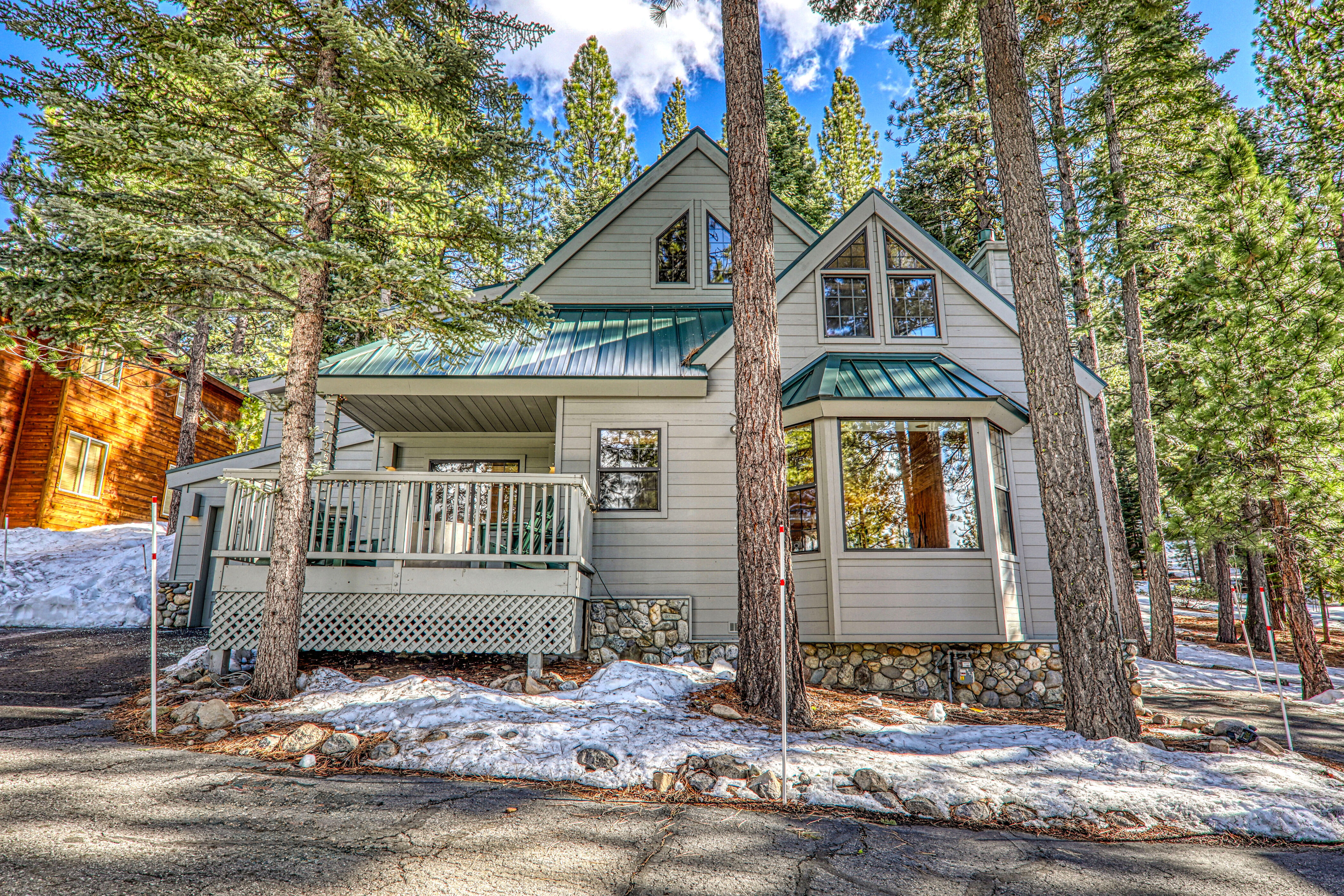 This charming, light-colored cabin-style home features a prominent A-frame roofline with metallic green accents and stone foundation skirting. The property includes a spacious elevated wooden deck and is nestled in a serene, wooded environment surrounded by tall pine trees and patches of remaining snow. It presents a cozy and inviting mountain aesthetic perfect for a vacation or rustic retreat.