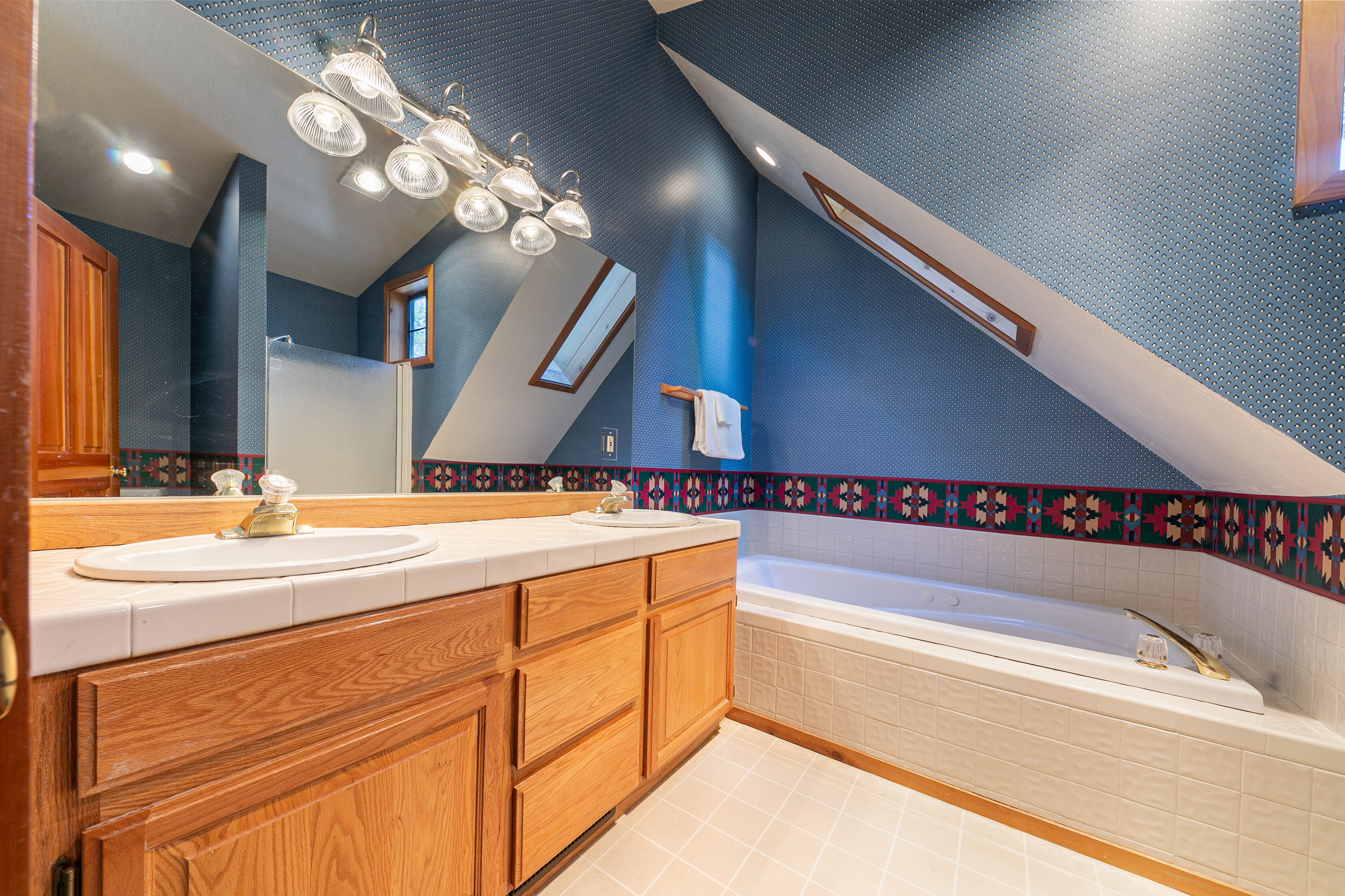 This primary bathroom features a dual-sink vanity with a tiled countertop and medium-toned wood cabinetry. The space includes a built-in jetted tub surrounded by white square tiles and is accented with blue wallpaper featuring a repetitive dot pattern and a colorful Southwestern-style border. The room is further characterized by a sloped ceiling and an angled skylight, creating a unique architectural aesthetic.