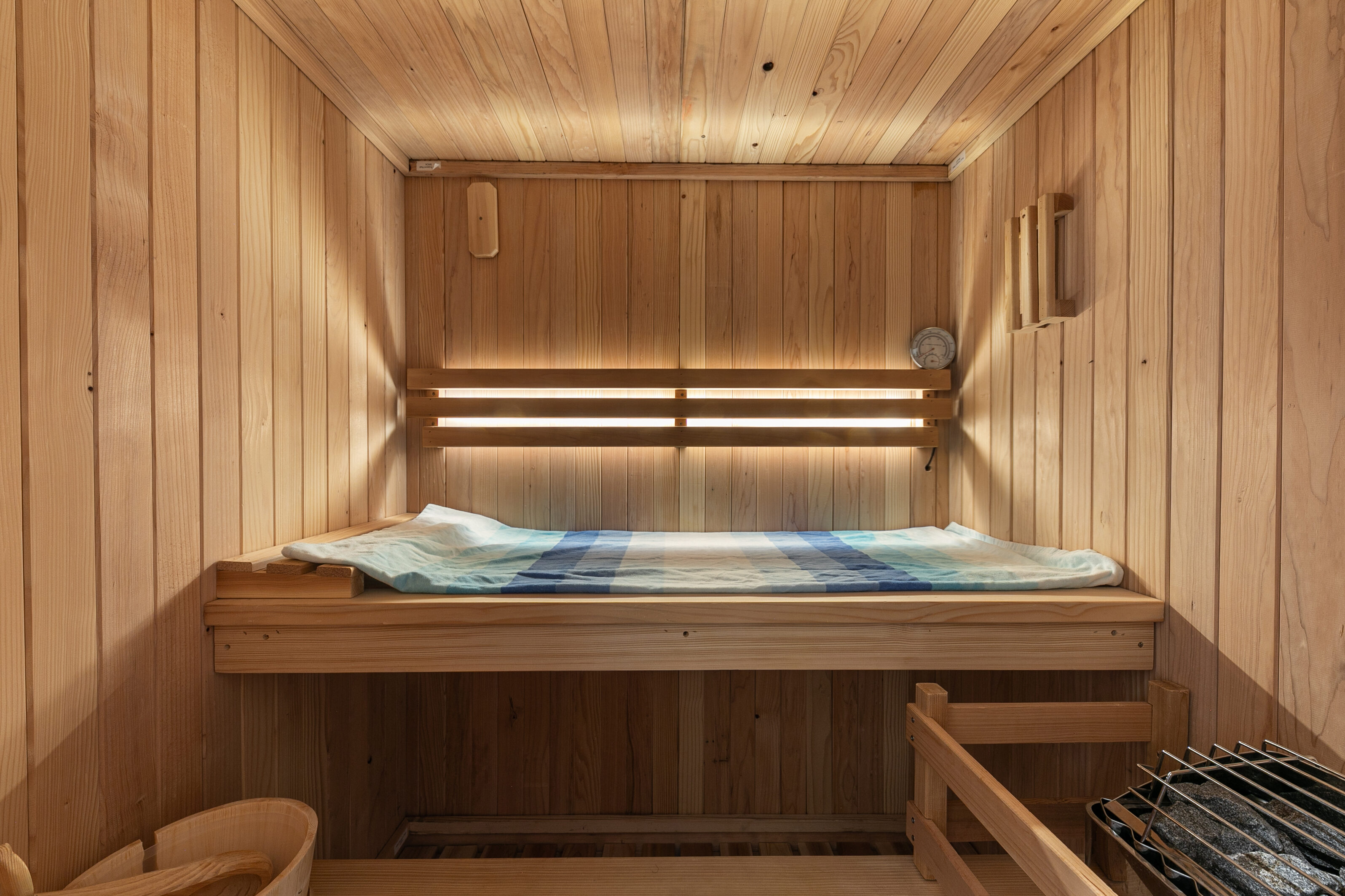 This is a custom-built, wood-paneled sauna featuring a tiered bench design with integrated cove lighting. The space is constructed from natural light-toned wood, creating a warm and relaxing atmosphere, and includes a built-in heater with rocks and a wall-mounted temperature gauge. It is a well-maintained, private wellness feature that adds significant appeal to a high-end luxury property.
