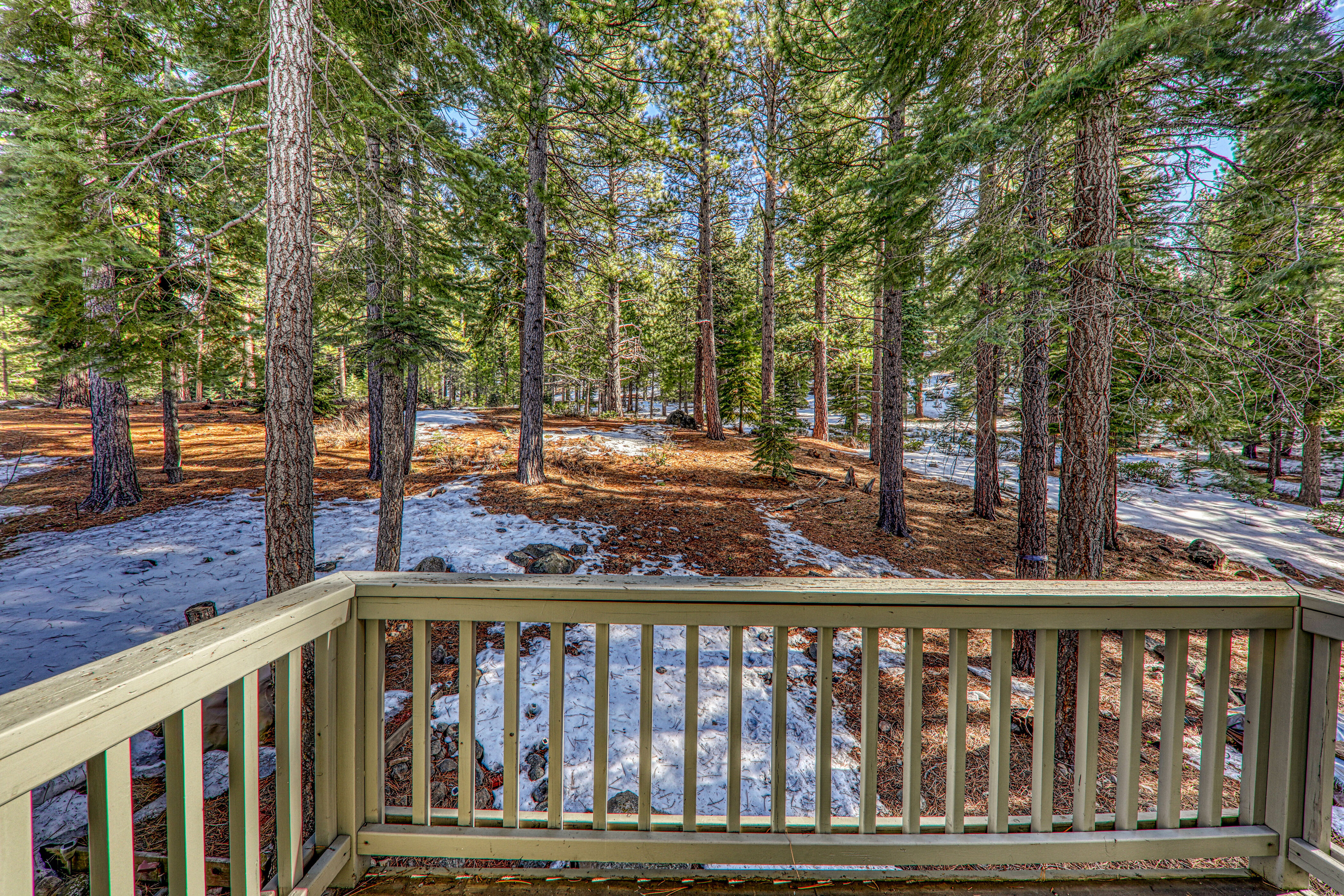 This image features a wooden deck balcony overlooking a tranquil, snow-dusted forest landscape. The simple railing provides a clear vantage point for enjoying the natural scenery and mature pine trees. It offers a serene and rustic outdoor retreat that enhances the property's connection to its wooded environment.