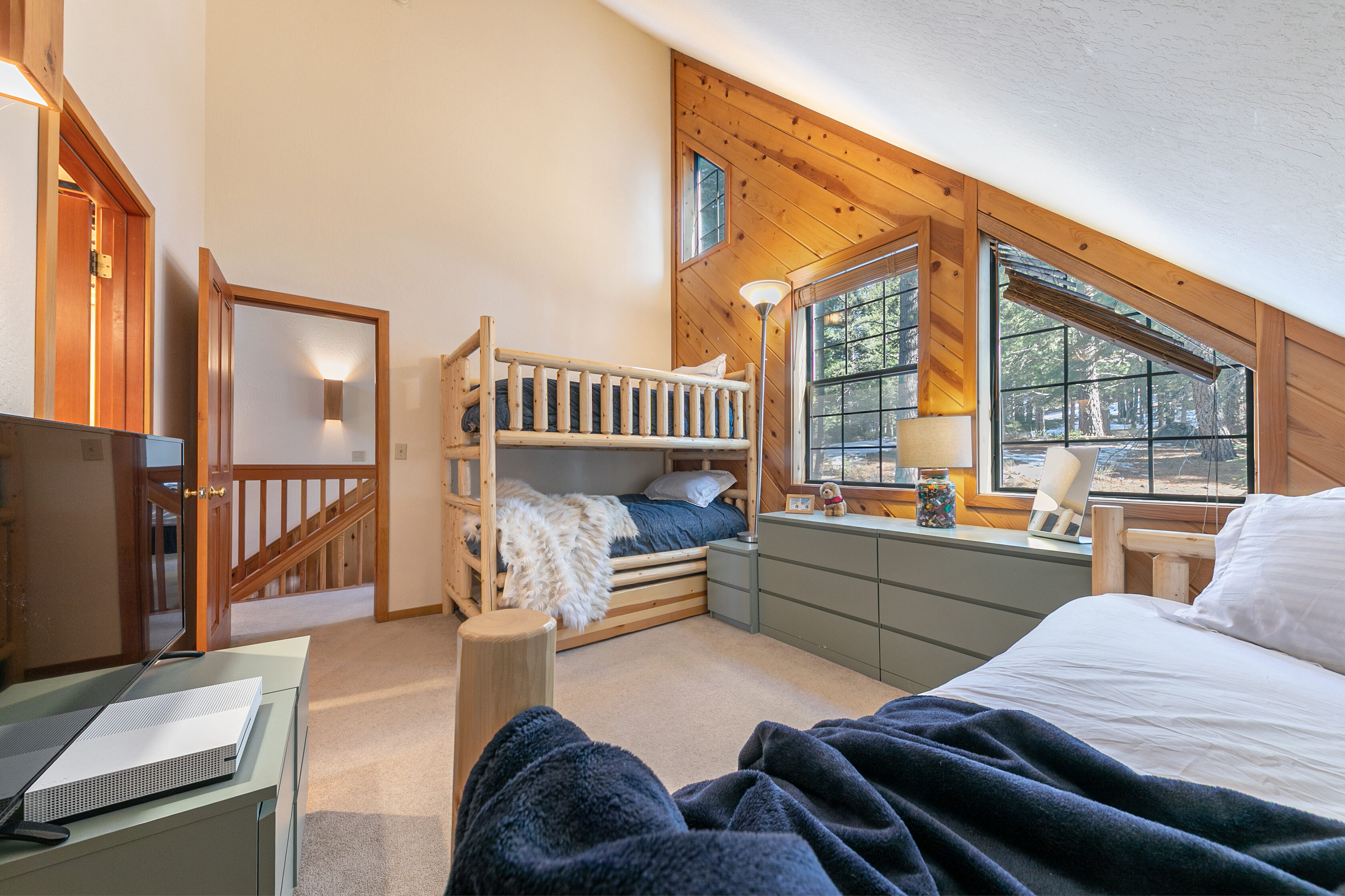 This cozy bedroom features rustic wooden-paneled walls and ceilings, giving it a warm, lodge-like aesthetic. It is furnished with a bunk bed, a convenient dresser, and a bed in the foreground, all set against a vaulted ceiling with large windows that bring in natural light. The room is well-appointed with functional storage and provides a comfortable atmosphere, highlighted by views of the trees outside.