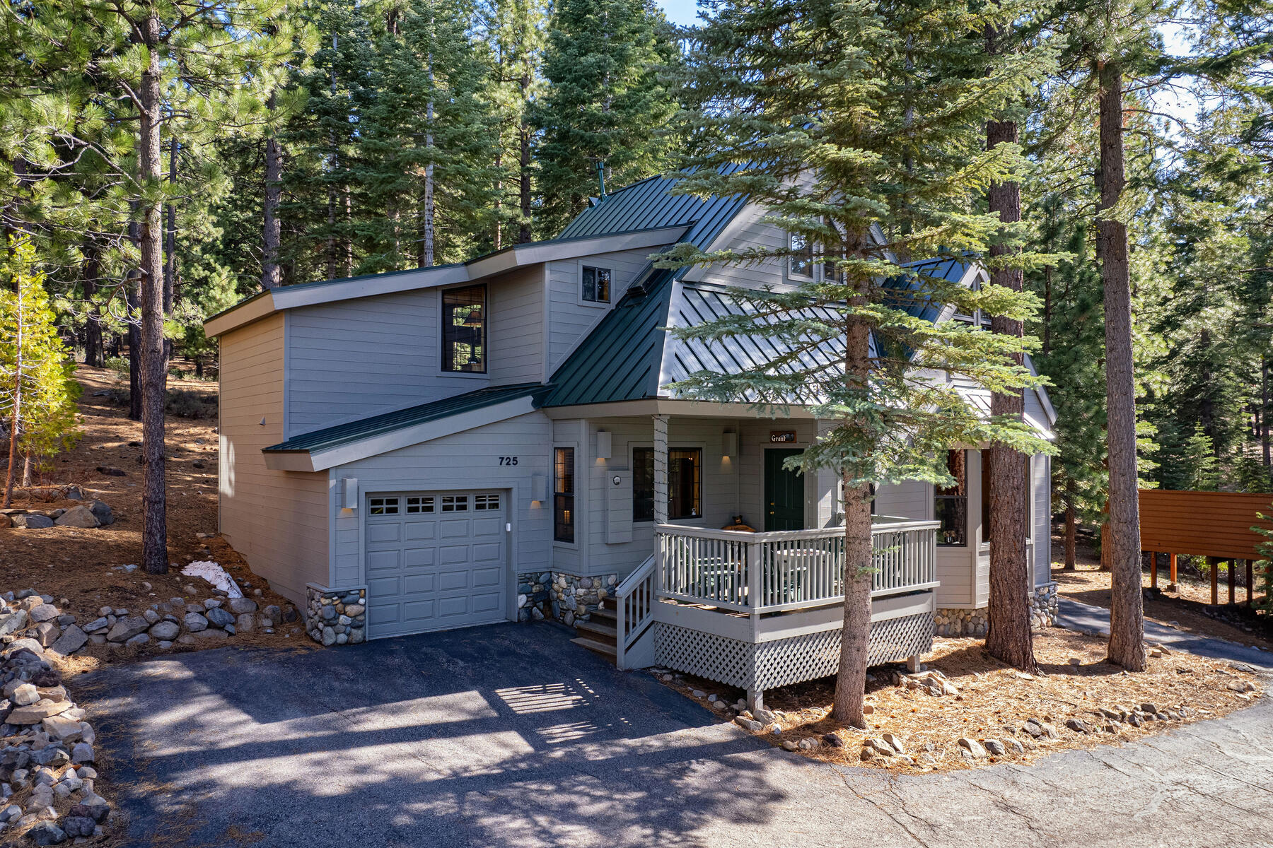 This charming, multi-level mountain home features a blend of light gray siding, natural rock accents, and a distinctive dark green metal roof. The property includes an attached garage and a welcoming front deck, all framed by a serene forest setting. Its cohesive design and rustic aesthetic create an inviting and peaceful curb appeal.