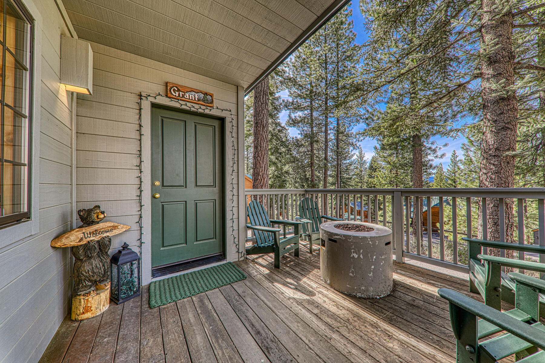 This cozy wooden deck features comfortable green Adirondack chairs surrounding a circular concrete fire pit, perfect for outdoor relaxation. The space includes a welcoming bear-themed statue near the entrance, enhancing the rustic, cabin-style aesthetic. Surrounded by tall pine trees, the deck offers a peaceful, scenic view perfect for enjoying the outdoors.