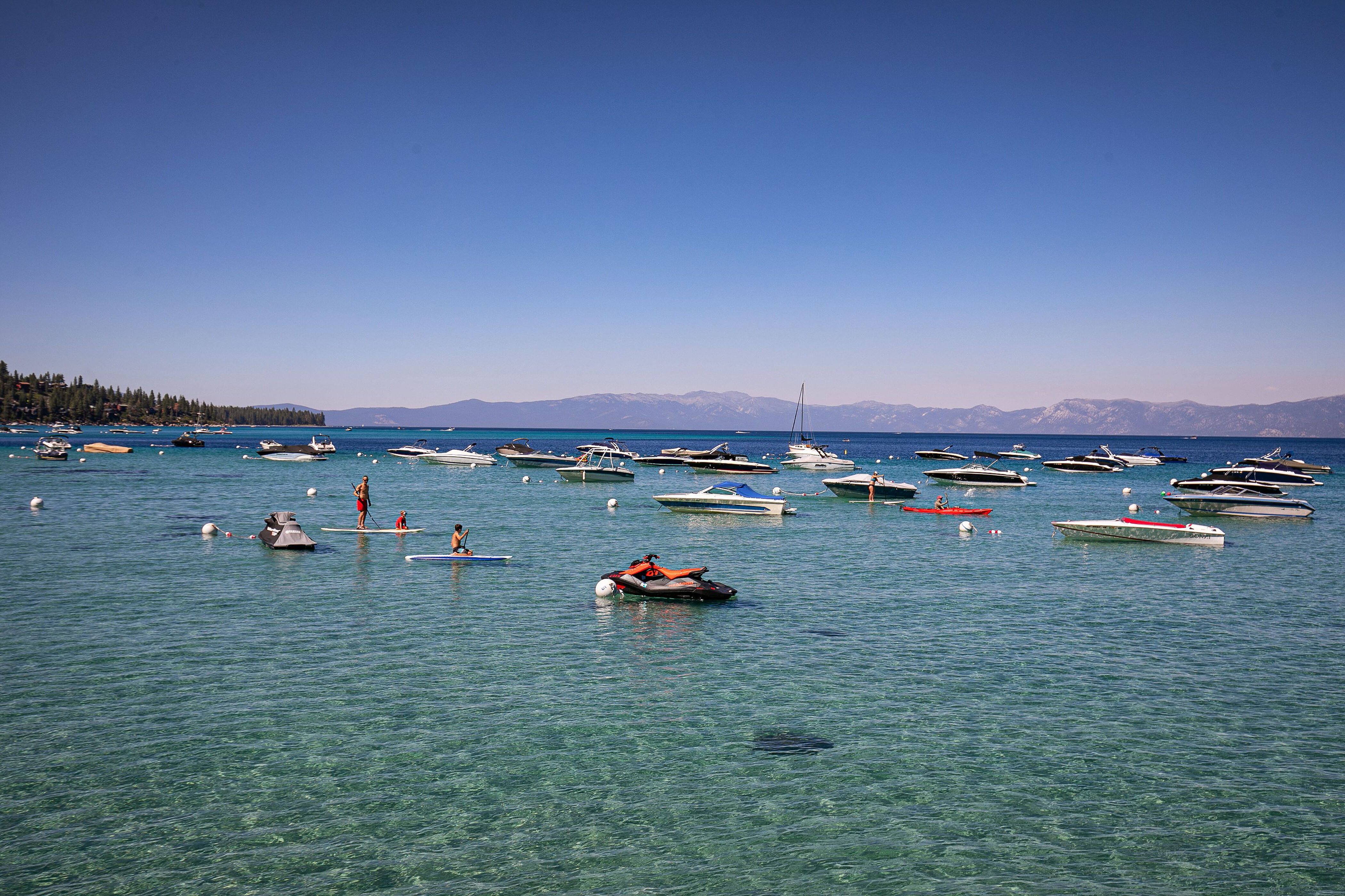 This image shows a scenic lakefront environment with clear, turquoise water and multiple recreational vessels, including boats, jet skis, and paddleboards. In the background, majestic mountains and dense evergreen forests frame the horizon, highlighting the natural beauty of the area. The clear blue sky and calm water suggest an idyllic outdoor lifestyle setting, making this an attractive feature for lakefront residential properties.