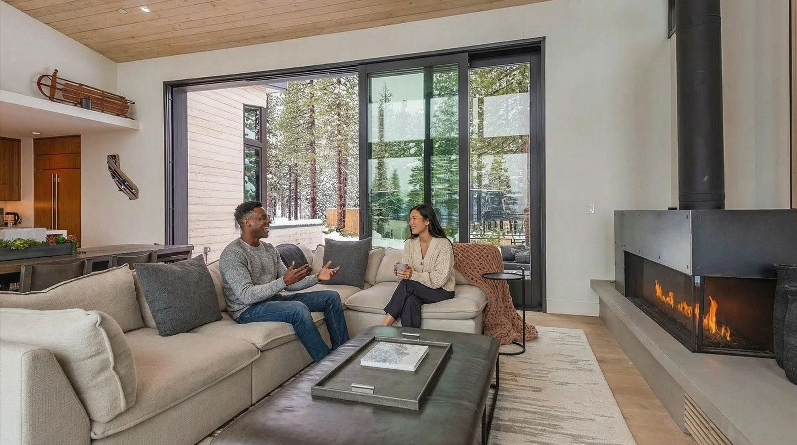 This is a bright and modern living room featuring a large sectional sofa, a fireplace, and expansive sliding glass doors that offer views of a snowy, tree-filled landscape. The interior design emphasizes natural light, with wood ceilings that complements the color scheme. The open-concept layout and comfortable seating arrangement creates an inviting atmosphere, perfect for relaxing and socializing.