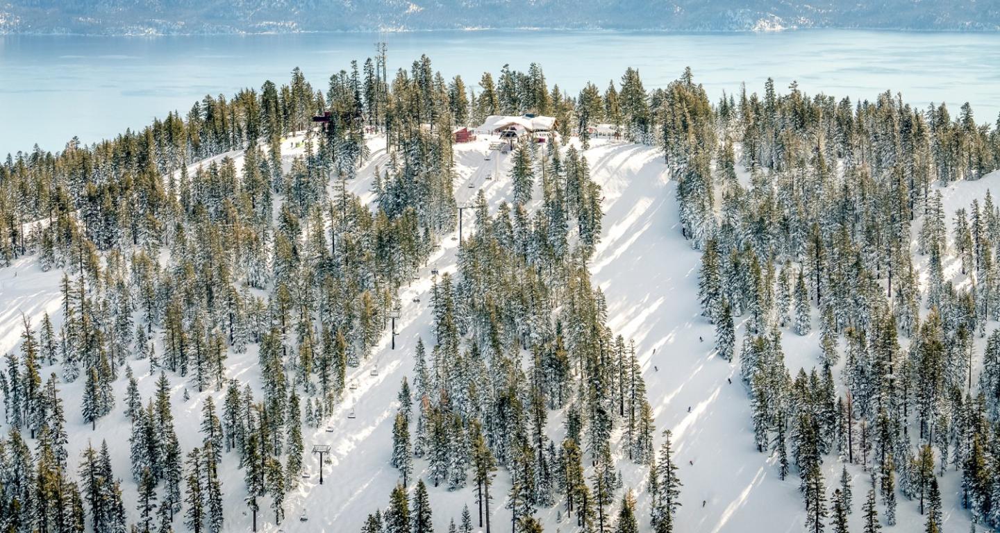 This aerial view showcases a ski resort nestled on a forested mountain slope, covered in snow. A building, likely a lodge or restaurant, is visible at the top of the mountain. The background features a pristine lake, contributing to a serene and picturesque setting, which creates an inviting atmosphere for potential buyers interested in vacation properties.