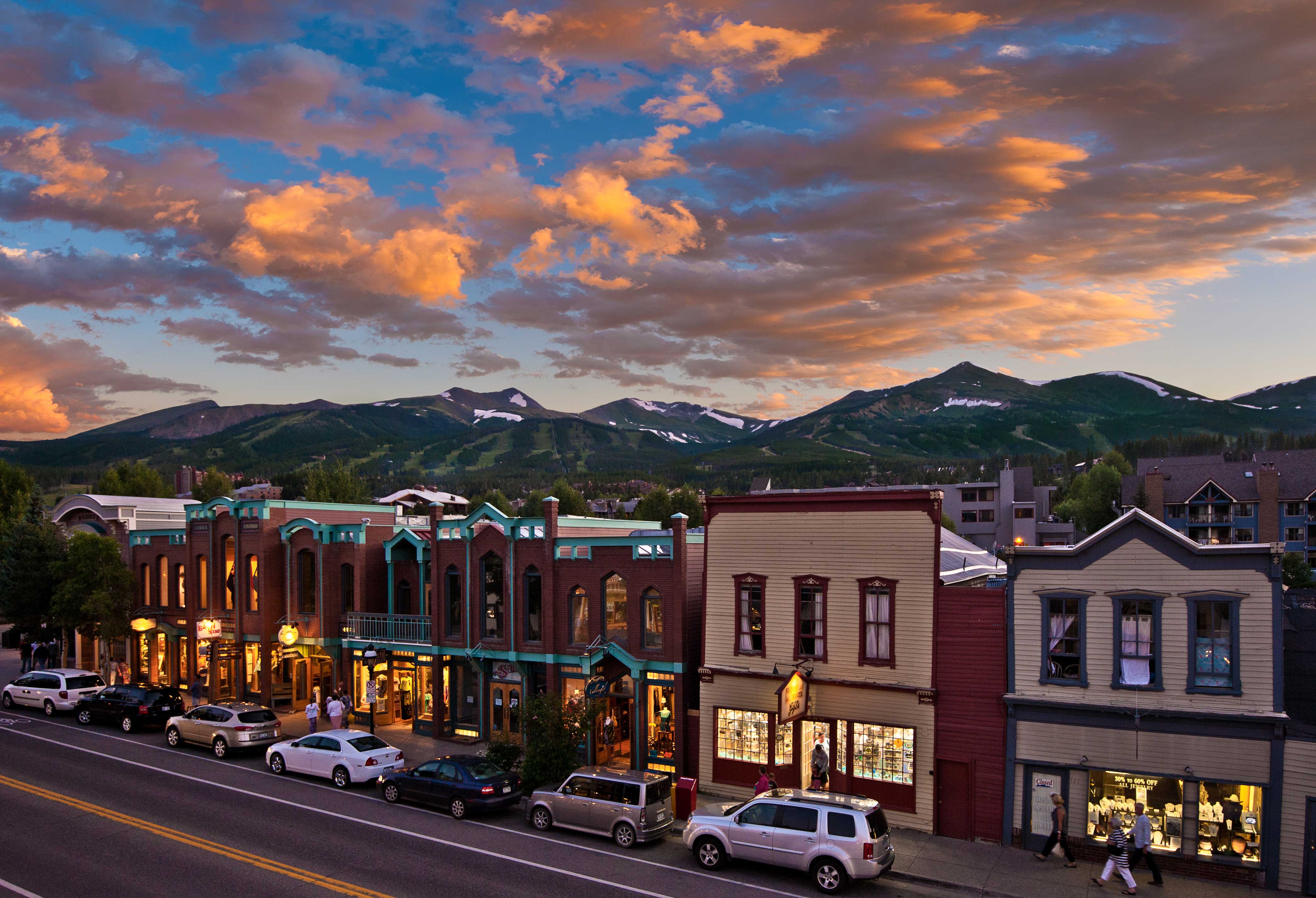 This image captures a charming street-level view of a historic downtown district, featuring traditional storefronts with ornate architectural details and storefront windows. The buildings are set against a breathtaking backdrop of snow-capped mountains under a vibrant, colorful sunset sky. This prime location suggests a highly desirable, walkable community with strong tourist and commercial appeal.