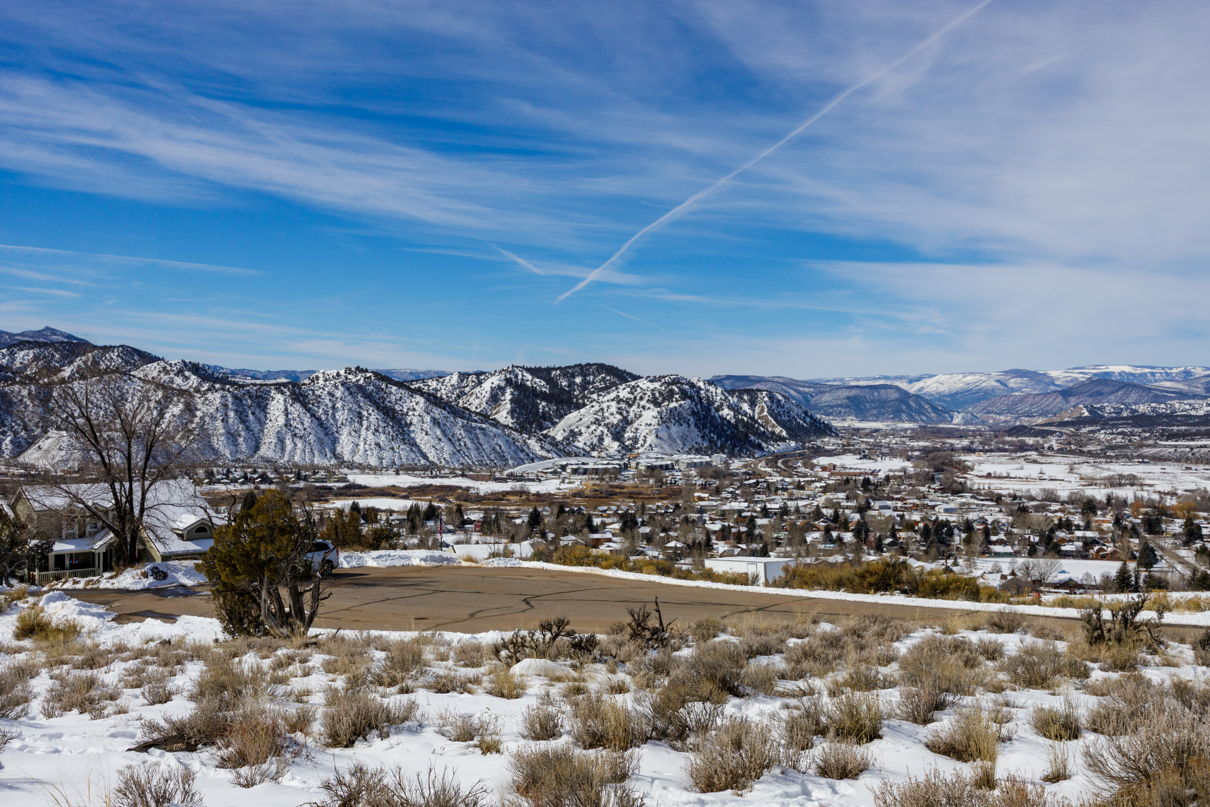 This aerial view showcases a residential area nestled in a valley surrounded by snow-capped mountains under a clear blue sky. Houses dot the landscape, with a mix of trees and open spaces creating a balanced and scenic environment. The image emphasizes the property's location within a peaceful and attractive setting.