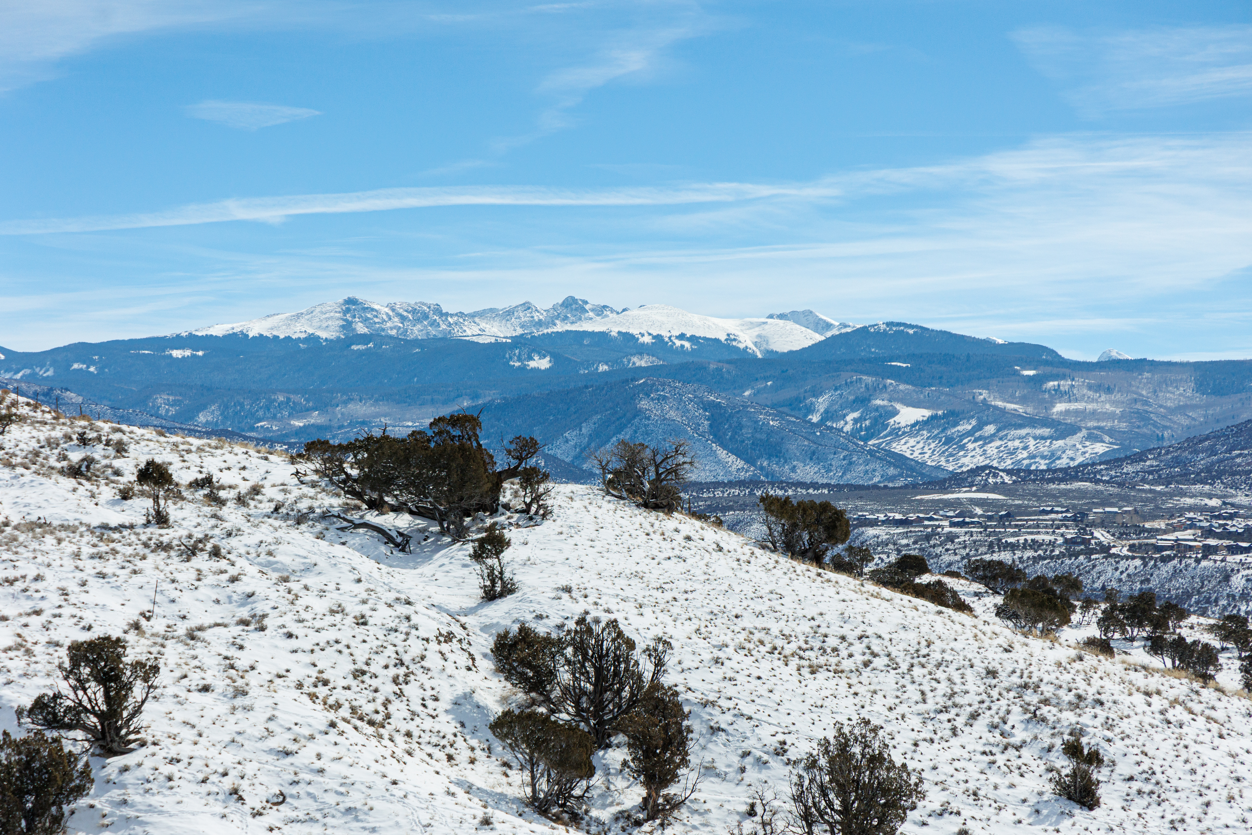 This aerial view showcases a snow-covered hillside dotted with trees, set against a backdrop of mountain ranges under a clear blue sky. The scene conveys a sense of serene isolation and natural beauty, appealing to buyers seeking a secluded property with picturesque views. The image emphasizes the vast, untapped potential of the land.