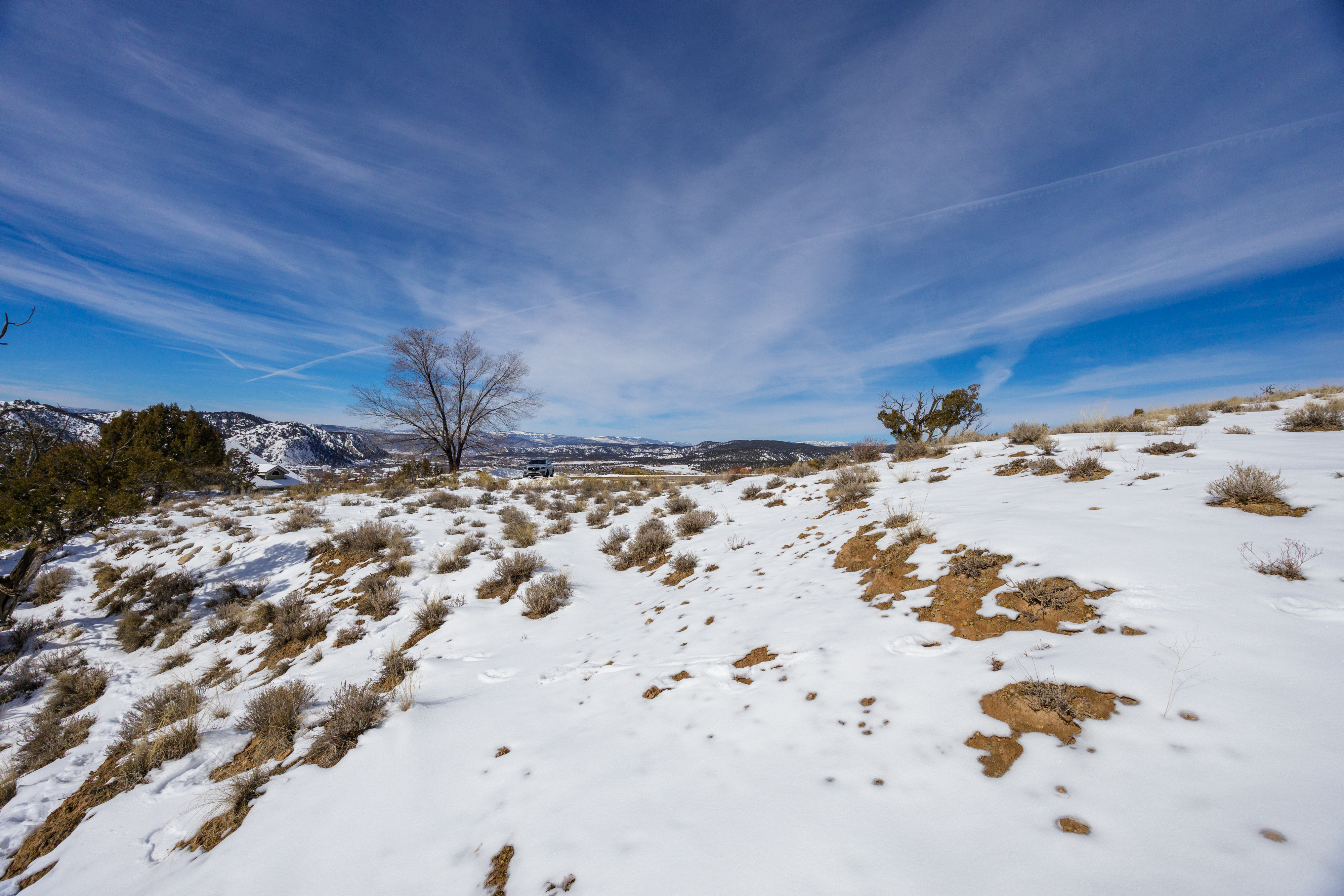 The image showcases a snow-covered yard or garden, featuring natural terrain with scattered vegetation. A clear blue sky with wispy clouds forms the backdrop, enhancing the openness of the landscape. The overall impression is of a serene and natural outdoor space, potentially highlighting the property's acreage and scenic views.