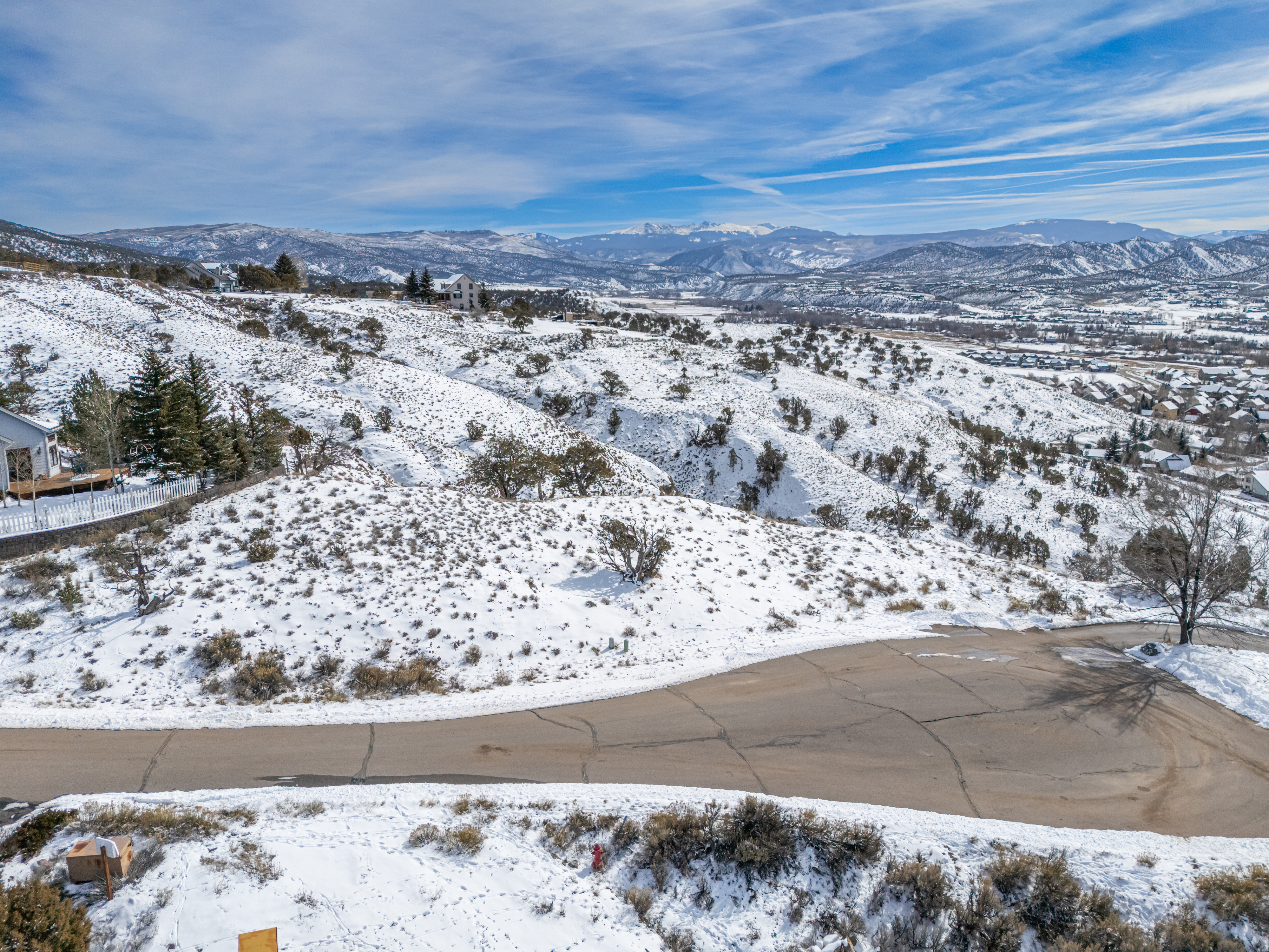 This aerial shot presents a snow-covered landscape featuring a residential area nestled amongst rolling hills. A paved road winds through the scene, while the backdrop showcases snow-capped mountains under a partly cloudy sky. This serene setting could appeal to buyers seeking a peaceful retreat with scenic views.