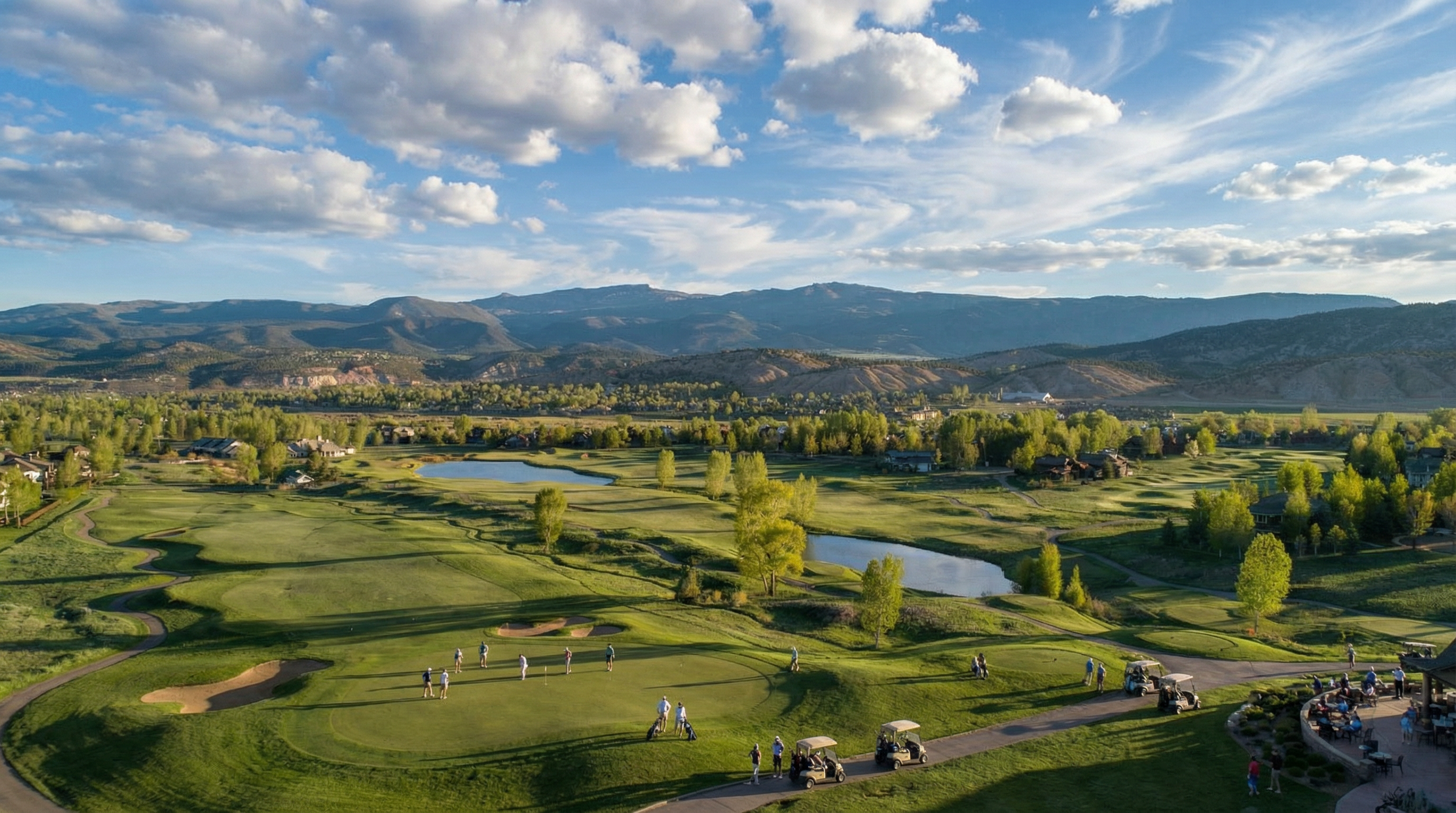 This aerial view showcases a meticulously maintained golf course community set against a backdrop of rolling mountains under a partly cloudy sky. Lush green fairways are punctuated by serene ponds and mature trees, highlighting the development's commitment to natural beauty and recreational amenities. A clubhouse or similar amenity is visible on the right side of the frame.