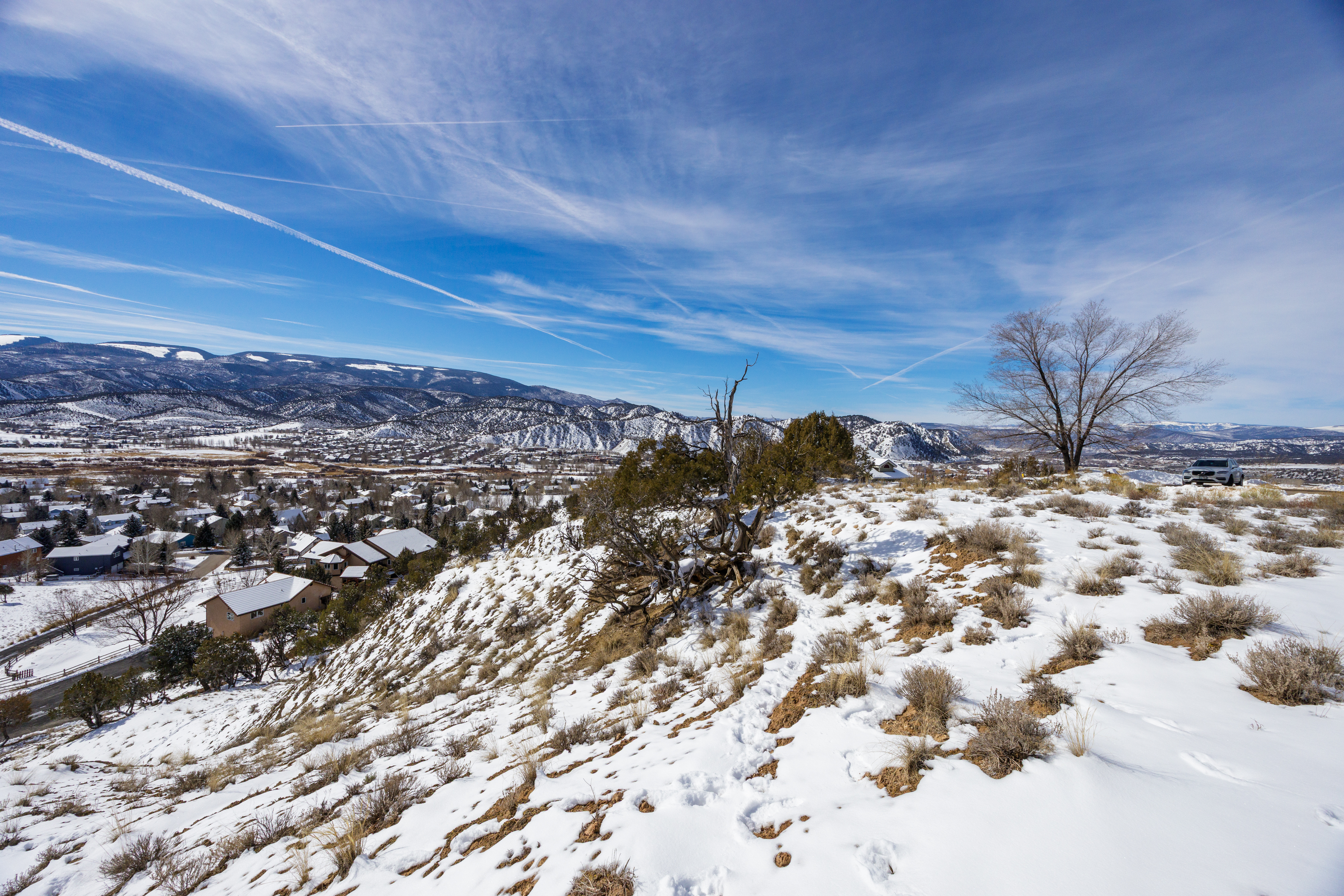 This aerial view showcases a residential area nestled at the foot of snow-capped mountains under a clear blue sky. The homes, some with visible snow-covered roofs, blend into the landscape, offering a sense of community and peace. A car is parked nearby, suggesting easy access and convenience for residents.