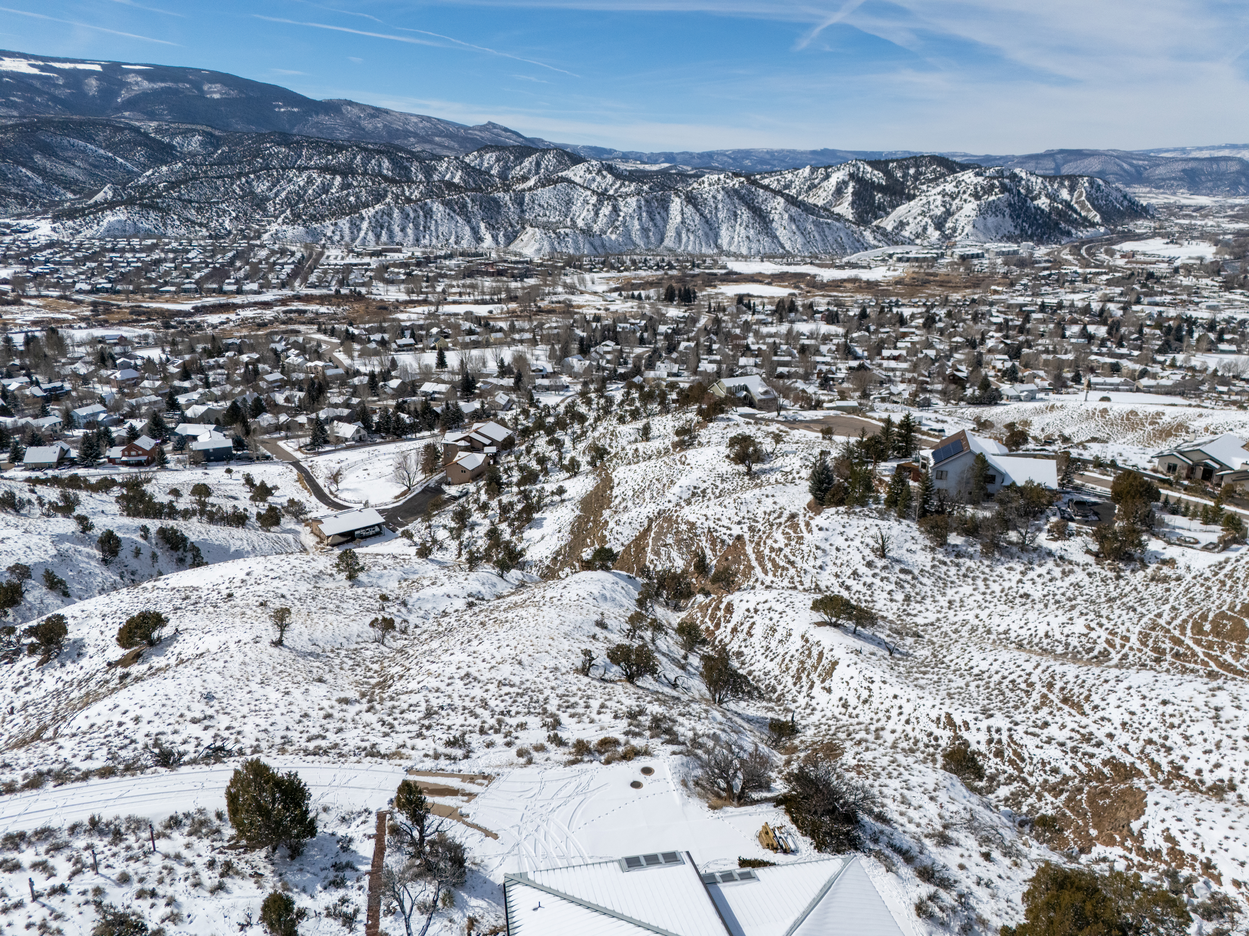 This aerial view showcases a snow-covered residential area nestled within a mountainous backdrop. The homes, varying in size and style, are blanketed in white, creating a serene winter landscape. The scene emphasizes the property's location within a scenic environment, highlighting its natural beauty and potential for a tranquil lifestyle.