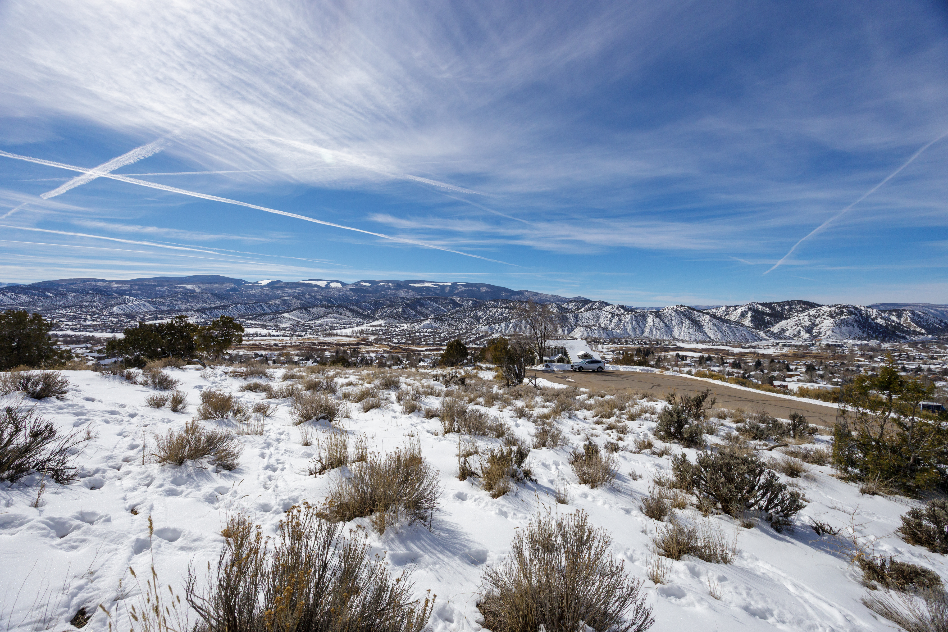 This image captures a scenic exterior view, likely showcasing a property nestled in a winter landscape. The foreground is characterized by snow-covered terrain and native shrubs, while the background reveals rolling hills and a town surrounded by mountains. A residential building is visible, suggesting a serene and picturesque living setting.