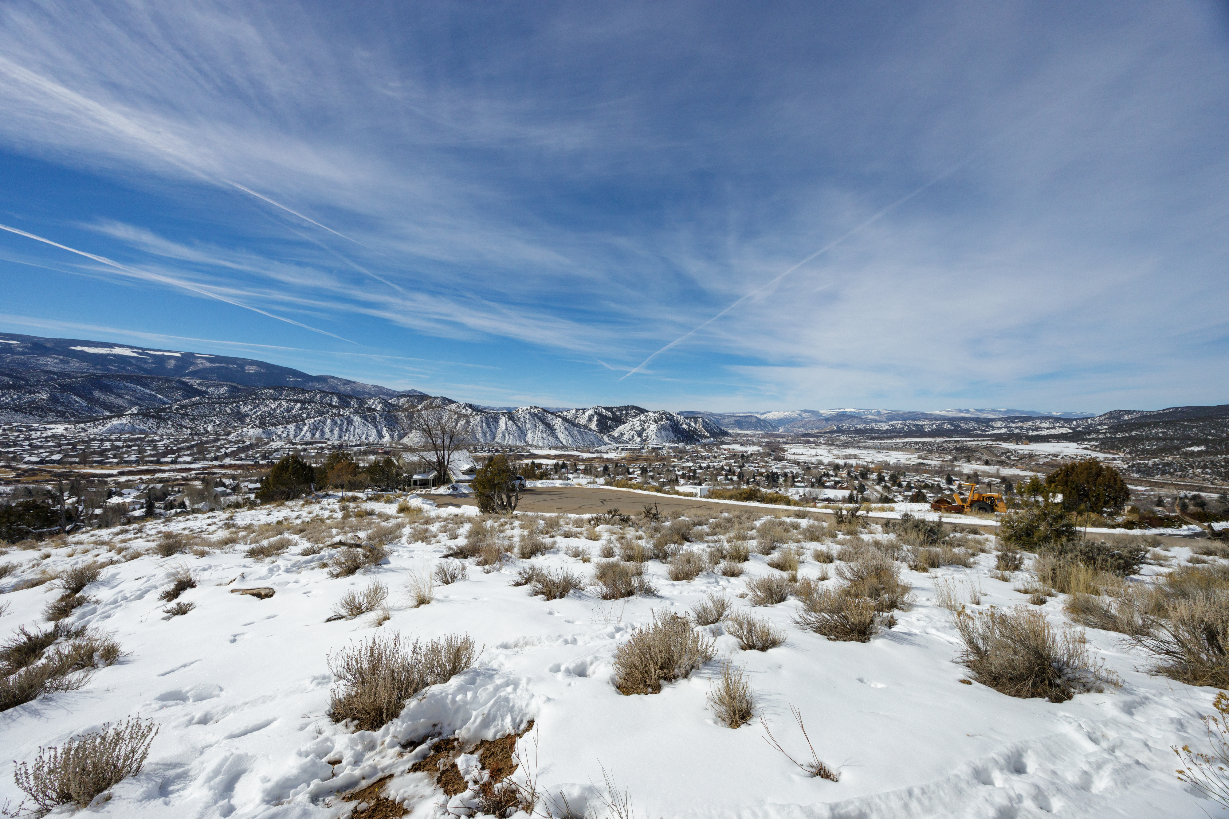 This aerial view showcases a scenic landscape with snow-covered terrain, shrubs, and scattered trees. In the distance, a town nestled amid rolling hills and mountains adds to the picturesque setting, creating a sense of serene living. The clear blue sky enhances the feeling of openness and tranquility, highlighting the potential of the location.
