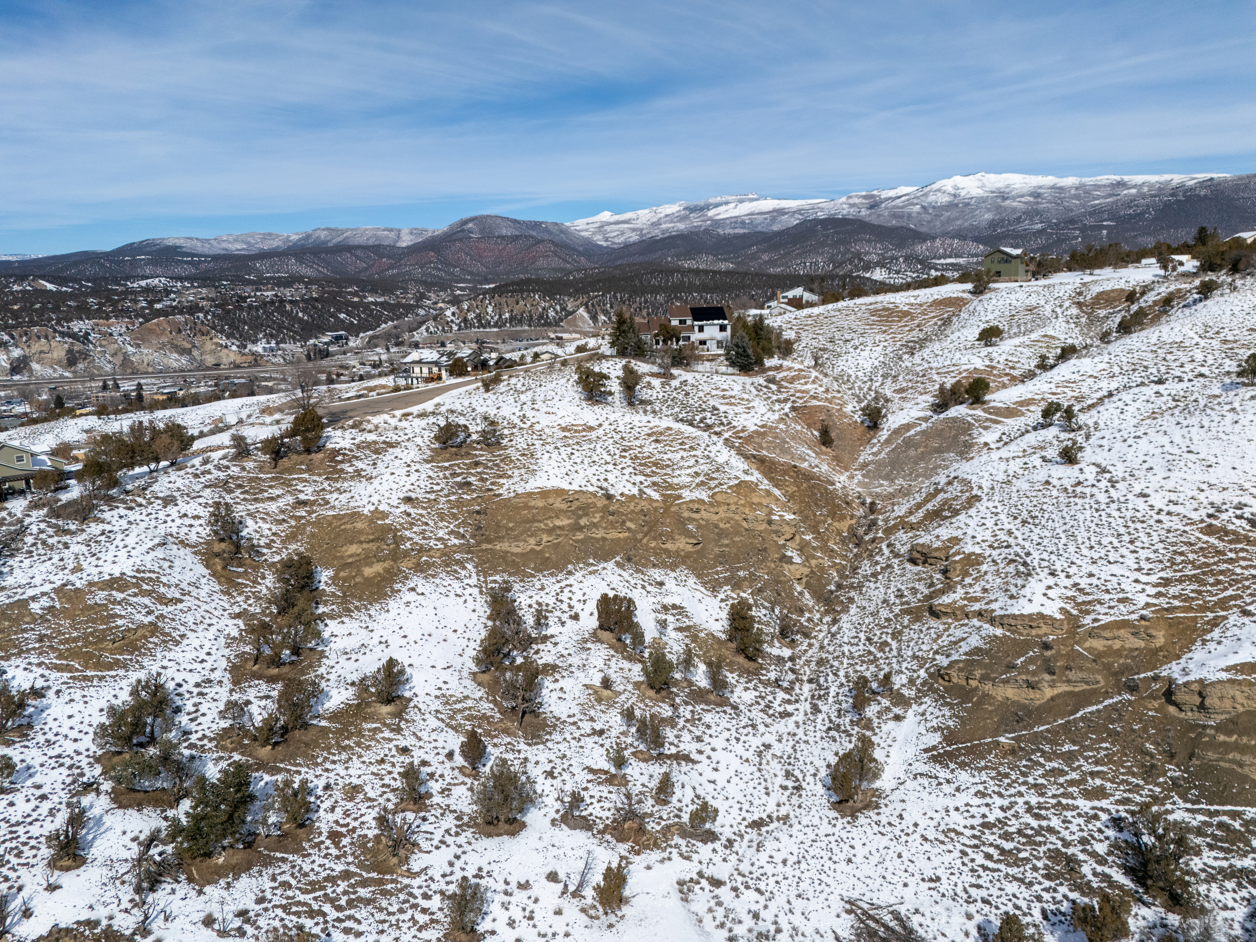 This aerial view showcases a property nestled in a snow-dusted, hilly landscape. A house with a modern design is visible on a slope, overlooking distant mountains and a town in the valley. The surrounding area features a mix of rocky terrain, scattered trees, and snow-covered areas, presenting a scenic and secluded setting.