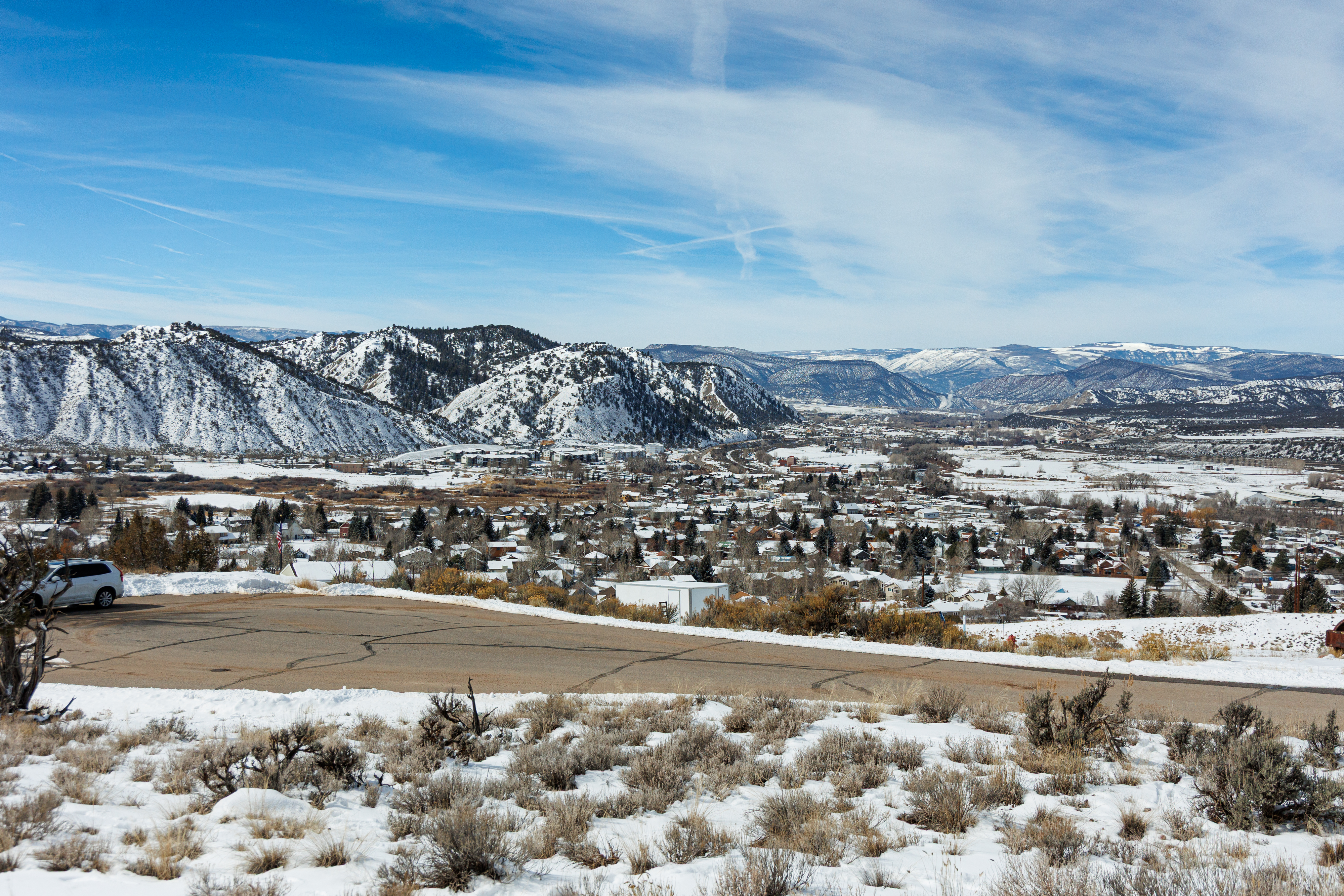 This image showcases an aerial view of a town nestled in a valley with snow-capped mountains in the background. The scene captures a mix of residential buildings, trees, and open spaces covered in snow. A paved road in the foreground adds depth and context to the setting which offers a picturesque winter landscape relevant to a real estate location.
