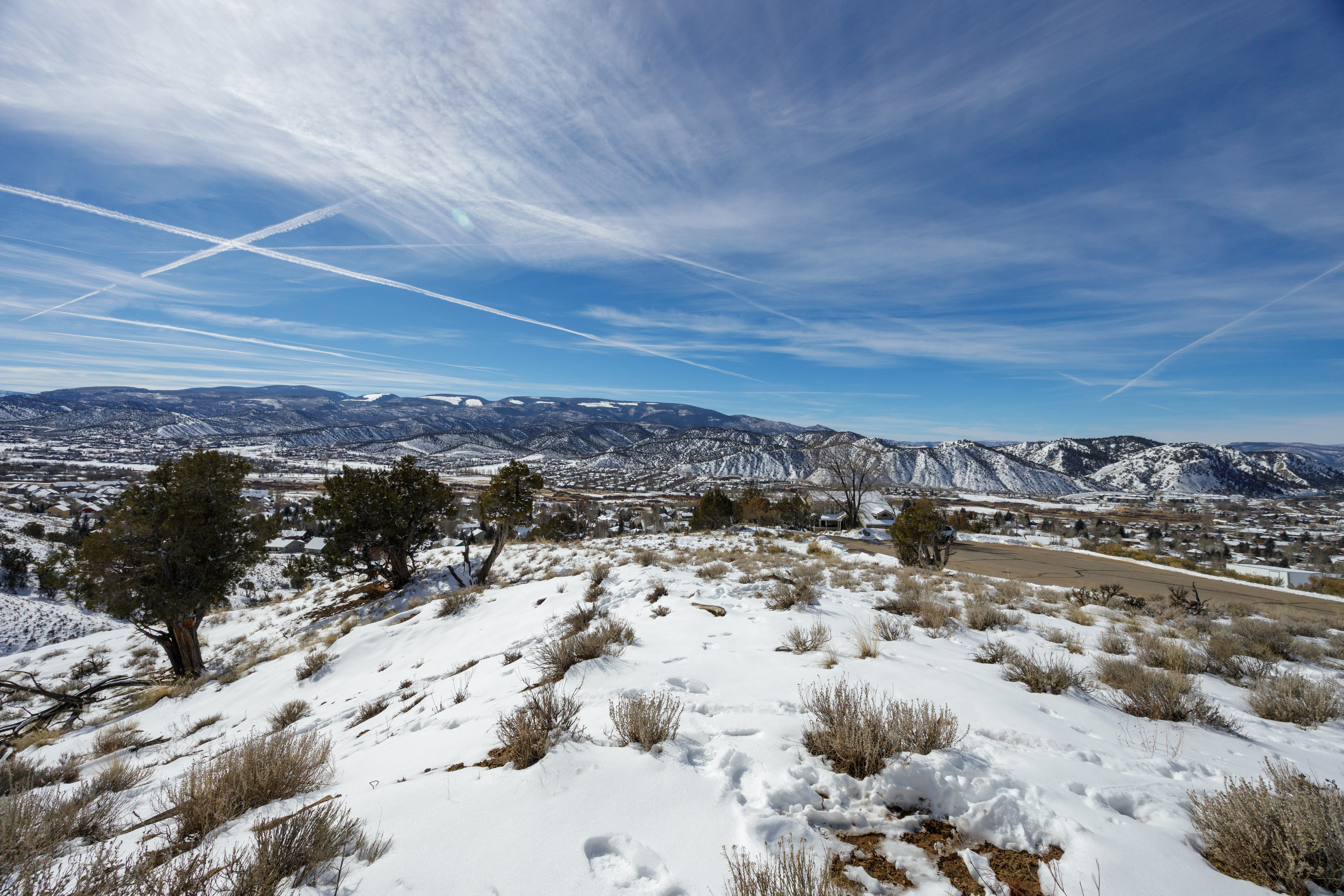This stunning aerial image showcases a winter landscape, with a snow-covered hillside leading to a panoramic view of a town nestled amidst mountains. Patches of vegetation break through the snow, offering visual interest and suggesting potential landscaping appeal. The expansive sky and the sense of elevation contribute to the property's desirable location and scenic views.