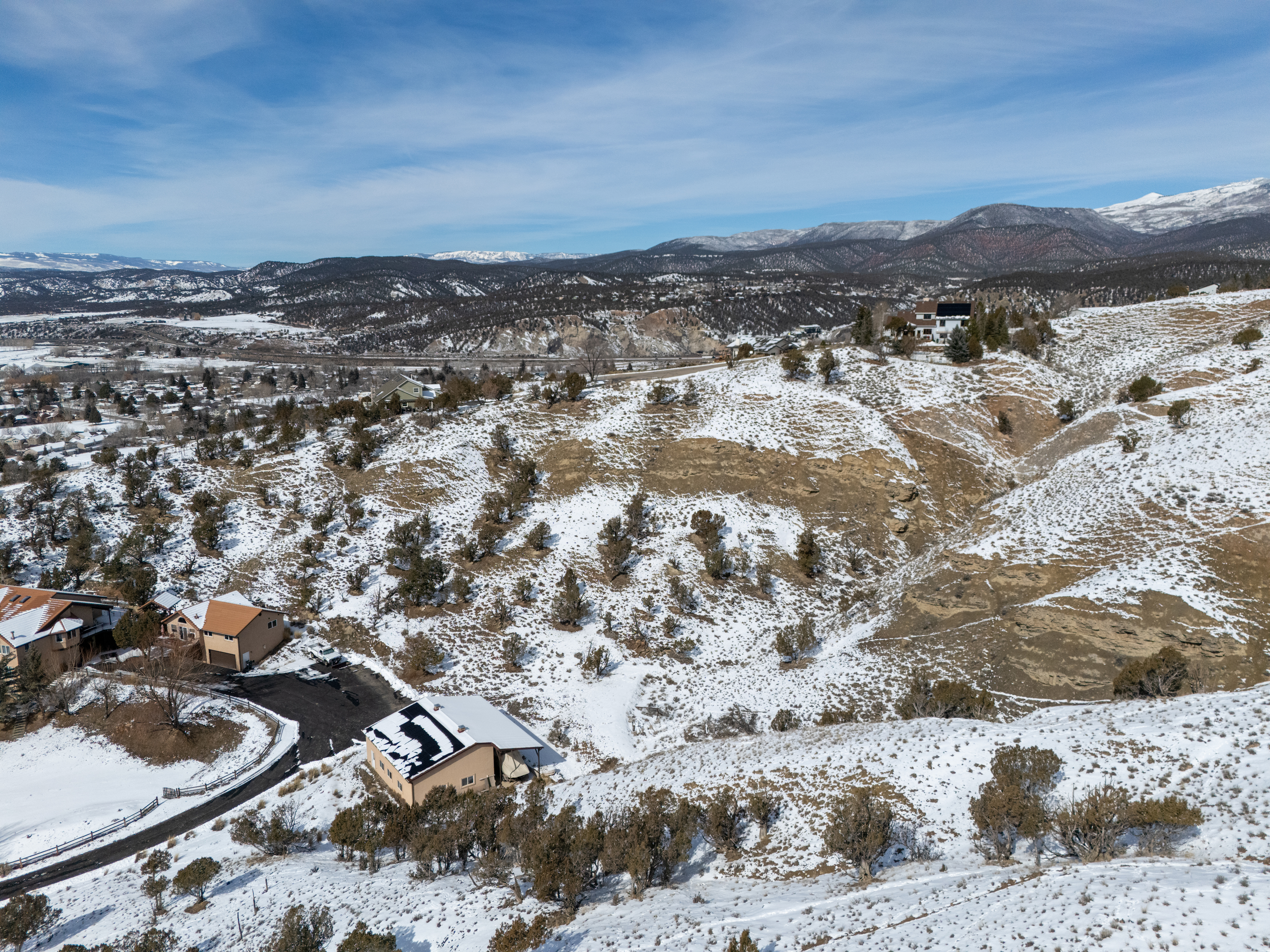 This is an aerial view of a residential area in a mountainous region partially covered in snow. Several houses are visible, nestled among trees and snowy patches, with mountains in the background. The image showcases the property's scenic location and the overall layout of the neighborhood.