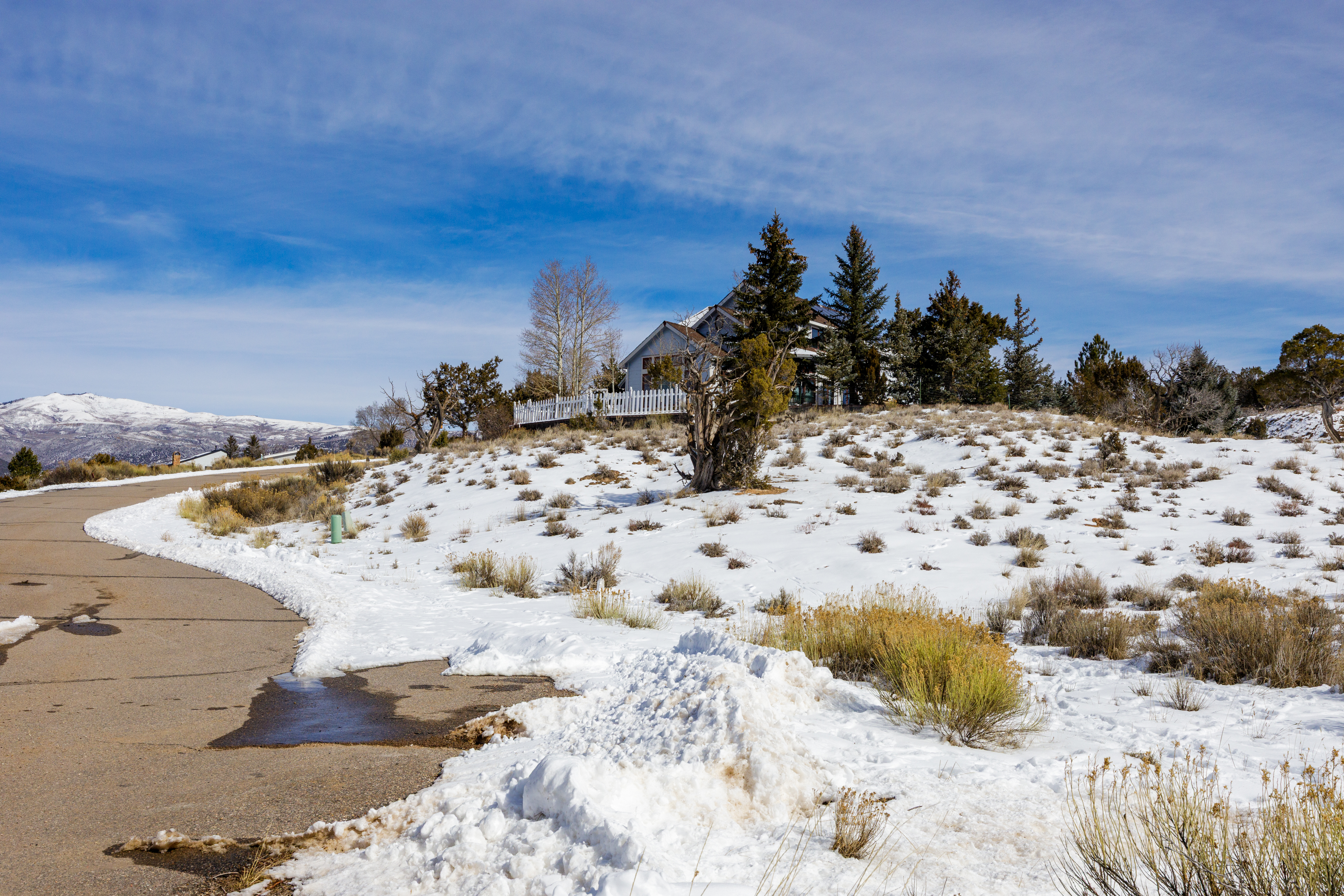 This is an exterior side view of a single-family home in a snowy, mountainous landscape. The house has a white picket fence and is surrounded by trees. The scene includes a paved road with patches of snow and the background features snow-capped mountains under a blue sky with light clouds, creating a serene and picturesque setting.