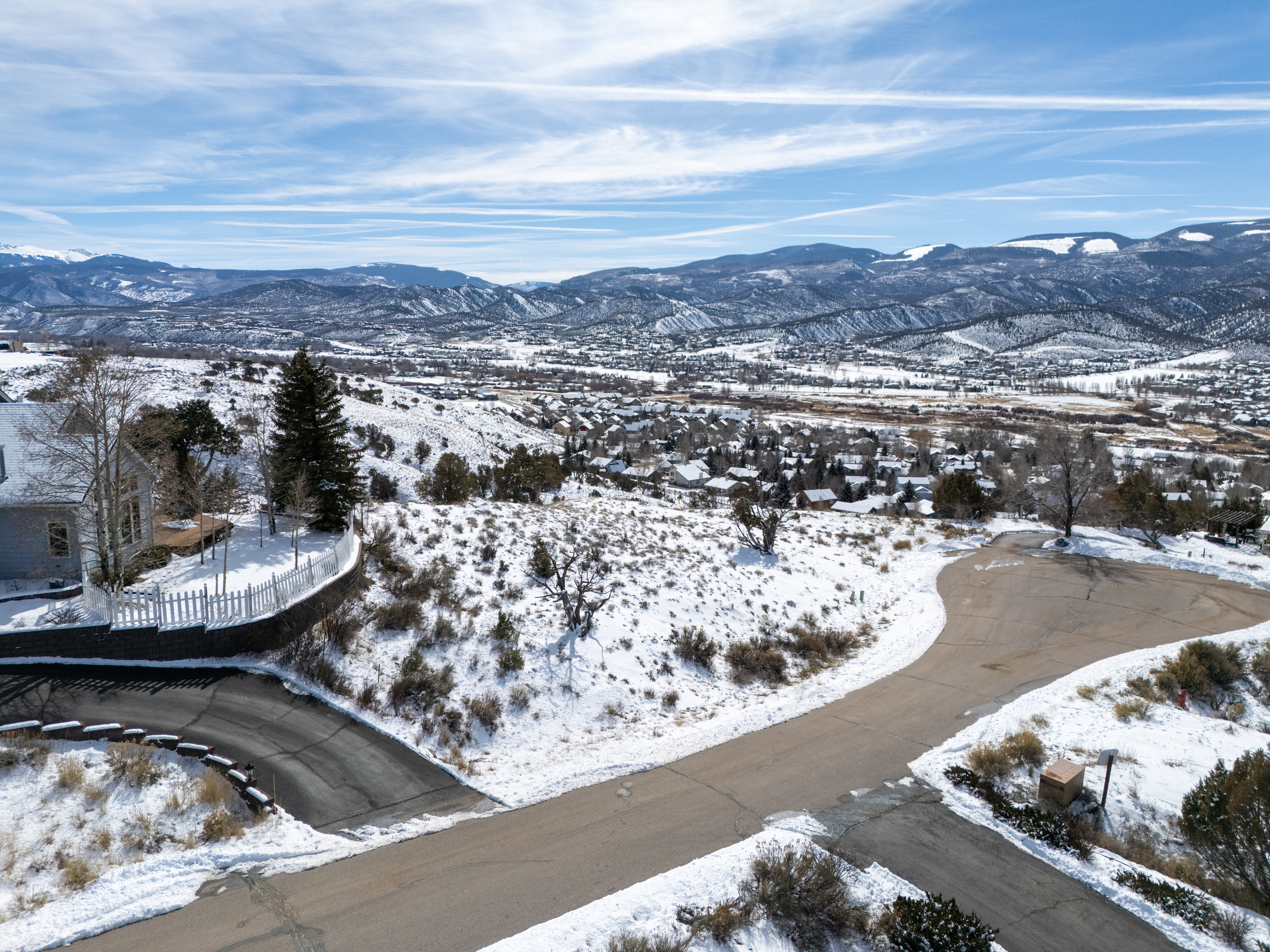 This aerial shot captures a scenic neighborhood nestled in a mountainous, snowy landscape. The winding roads and snow-covered houses present a serene winter view. The home is positioned on elevated land, which potentially boasts impressive mountain views.