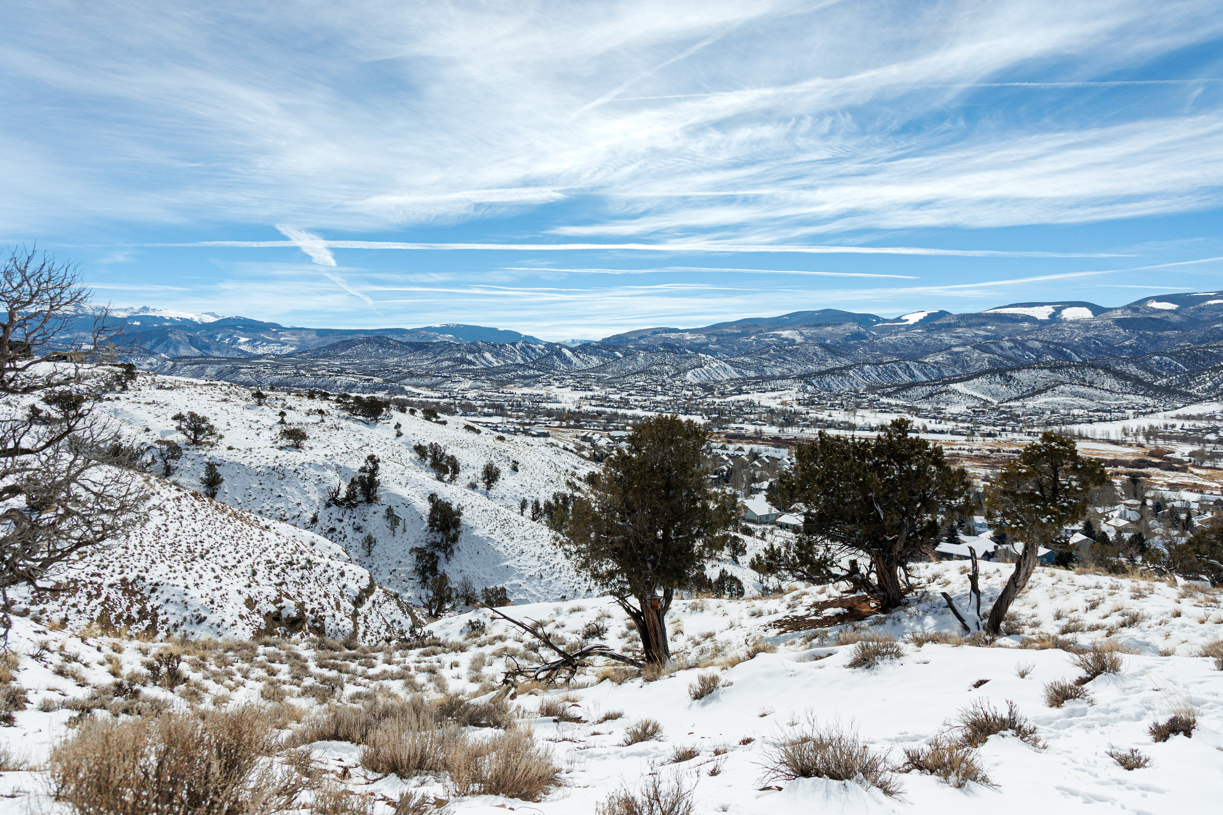 This is an aerial view of a snow-covered mountain landscape. The image captures a valley with scattered houses and a backdrop of mountains under a partially cloudy sky. The scene evokes a sense of serenity and isolation, highlighting the natural beauty of the area.