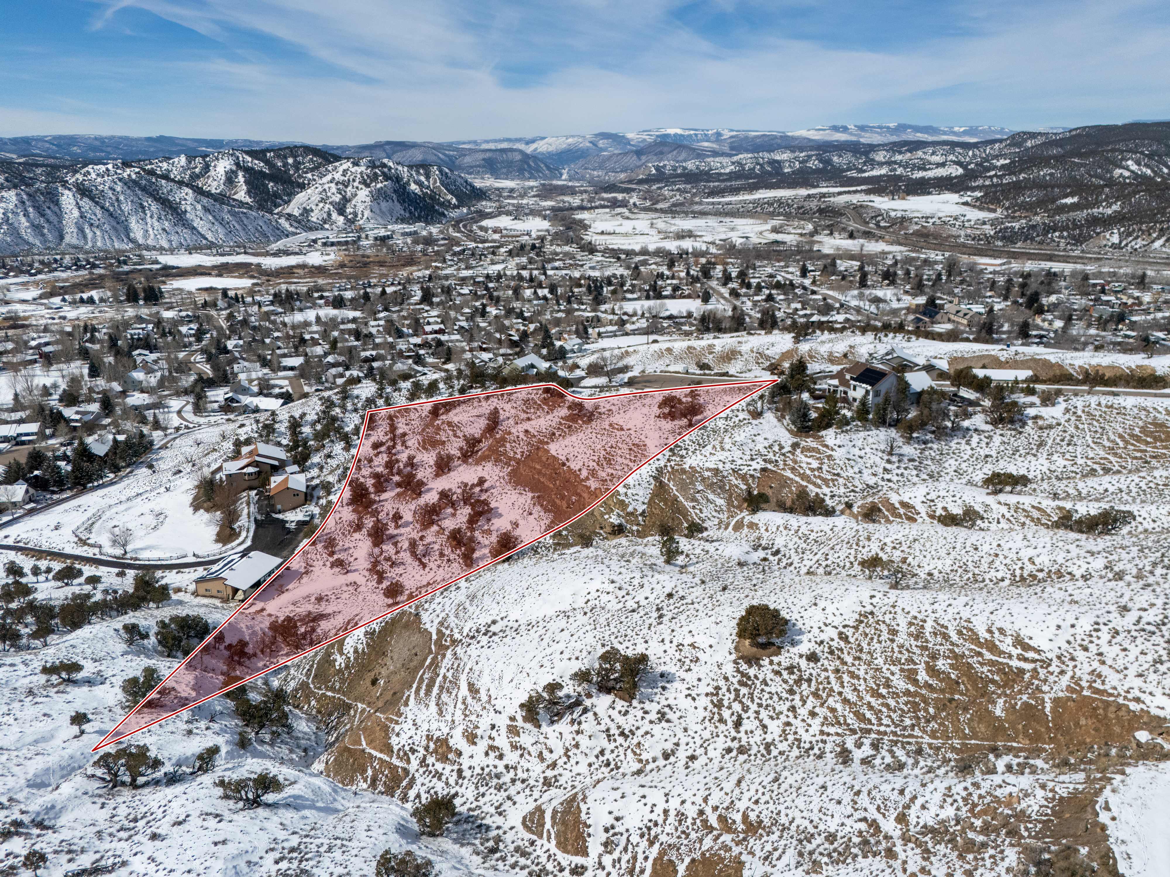 This aerial view showcases a large plot of land, possibly a property for sale, outlined in red. The land features some vegetation and snowy patches, set against a backdrop of a town and distant mountains. The image provides a sense of location and potential development opportunities.