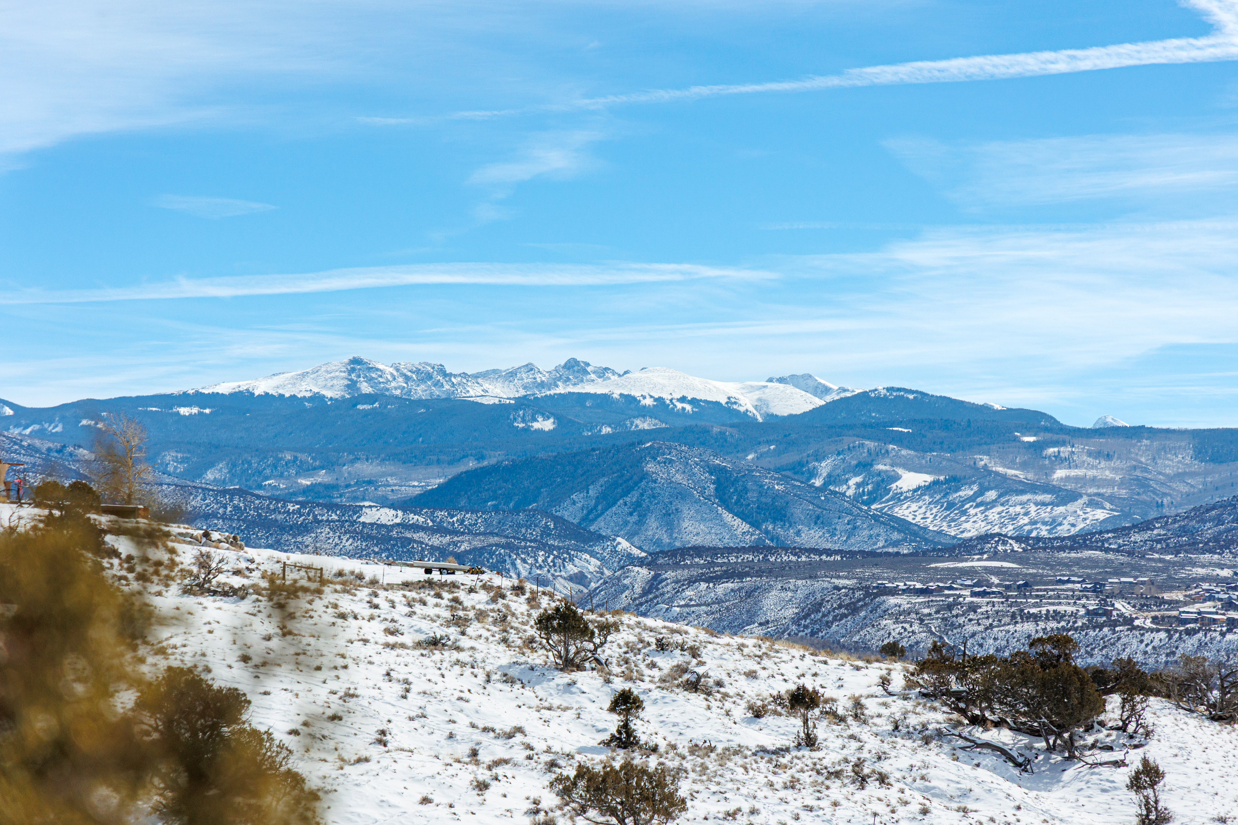 This aerial view showcases a snow-covered landscape with mountains in the background. The image captures the natural beauty and expansive scenery surrounding a property, emphasizing its location and potential for outdoor enjoyment. The presence of a neighborhood in the distance hints at accessibility and proximity to amenities.