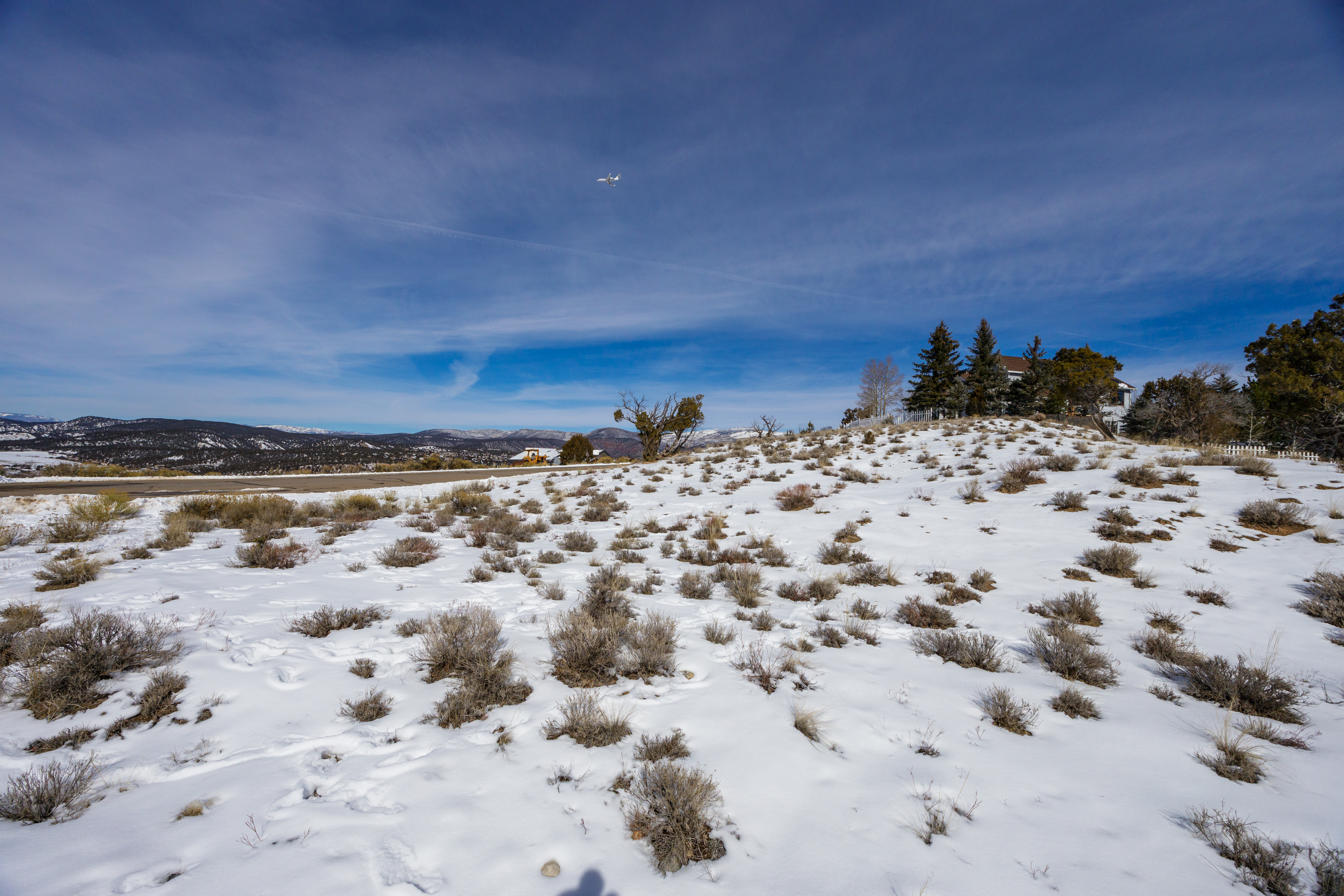 This image showcases the front exterior of a home set atop a snow-covered hill. The property features mature trees and possibly a white picket fence. An open landscape surrounds the house, providing a panoramic view, even extending to an airplane crossing overhead.
