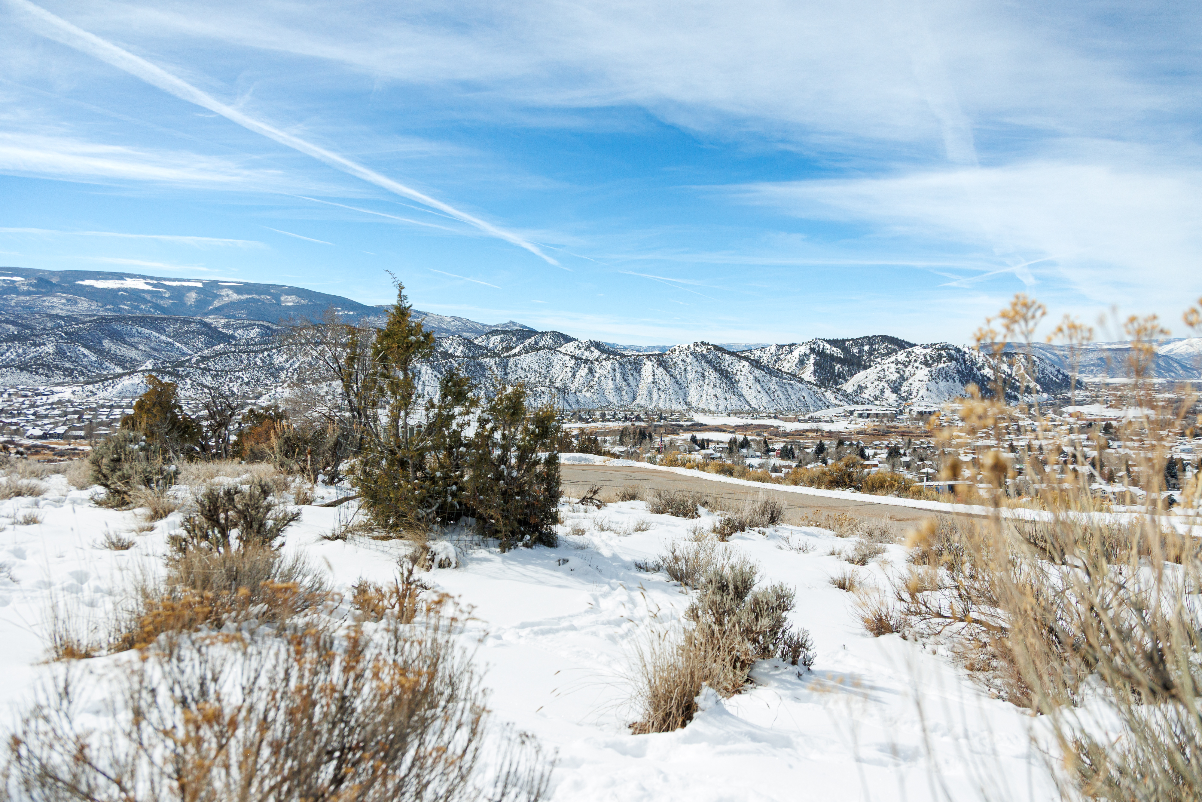 This aerial view showcases a snowy landscape with mountains in the background and a town nestled within the valley. The foreground features snow-covered terrain with scattered bushes and a road leading towards the town. The scene presents a serene and scenic environment, emphasizing the natural beauty surrounding the residential area.