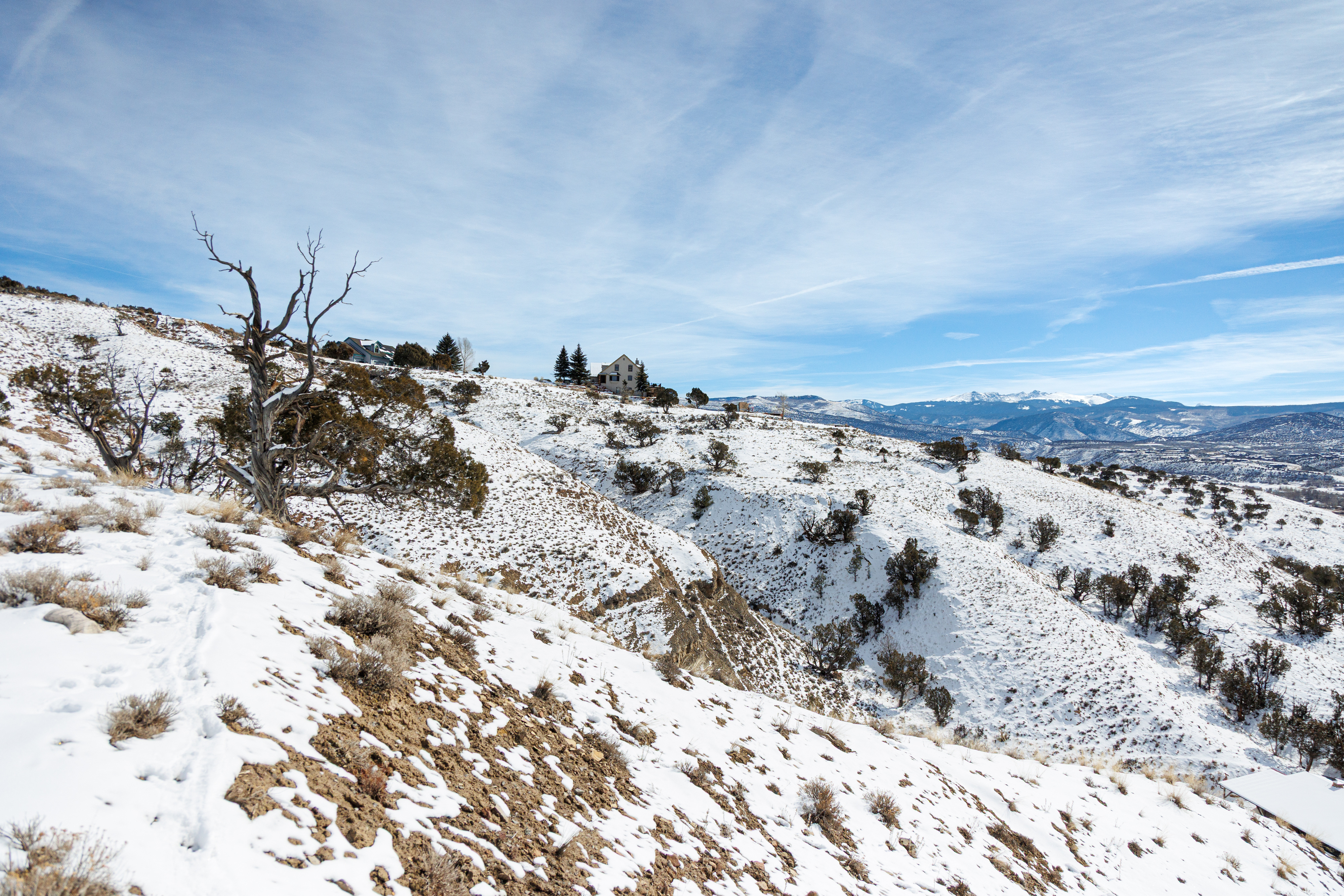 This image showcases a snow-covered landscape with scattered trees and shrubs, suggesting a spacious yard. In the background, a house is visible on a hill, potentially indicating a scenic location. The presence of snow and the distant mountains evoke a sense of tranquility and natural beauty, making it an attractive feature for prospective buyers seeking a peaceful environment.