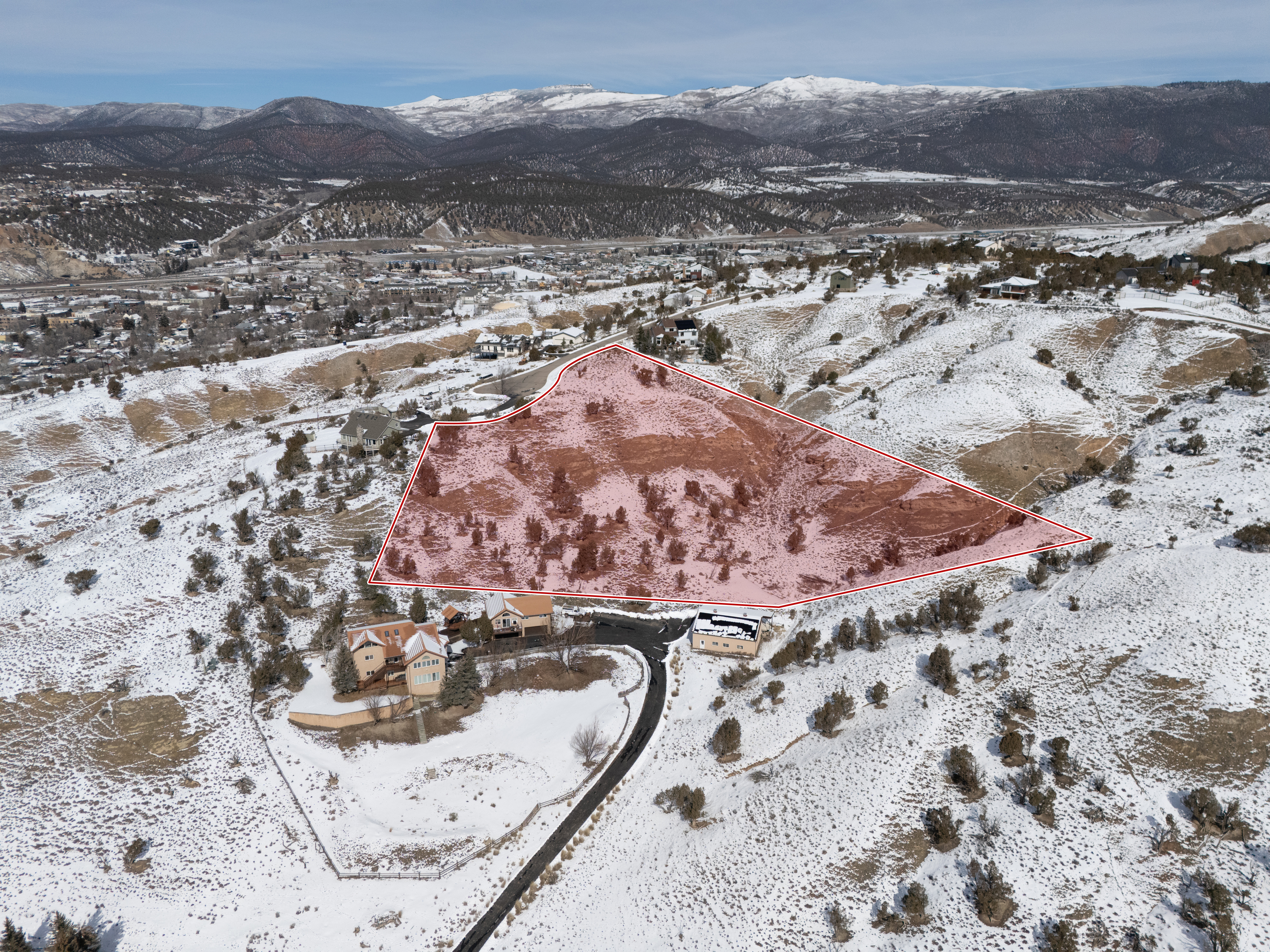 This aerial view showcases a parcel of land, possibly a building lot, highlighted with a faint pink overlay within a red border. The surrounding landscape is a mix of residential properties, snow-covered ground, and distant mountains. The image suggests a serene, elevated location with potential for a custom-built home.