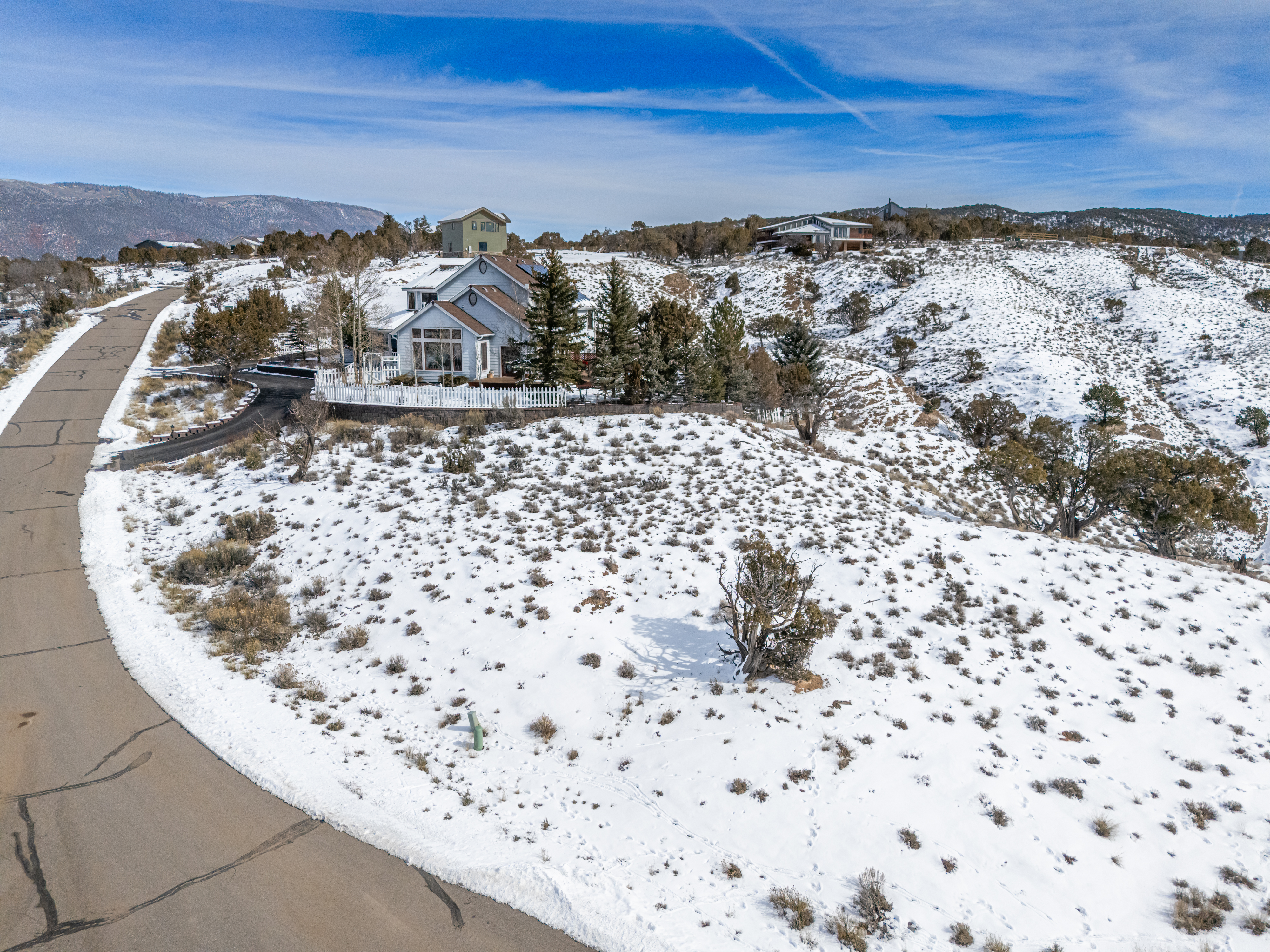 This aerial view showcases a hillside home nestled amongst snow-covered grounds and varied vegetation. A winding road leads up to the house, adding to the secluded and peaceful atmosphere. Other homes can be seen nestled into the hill in the distance, suggesting a community that enjoys elevated scenic views.