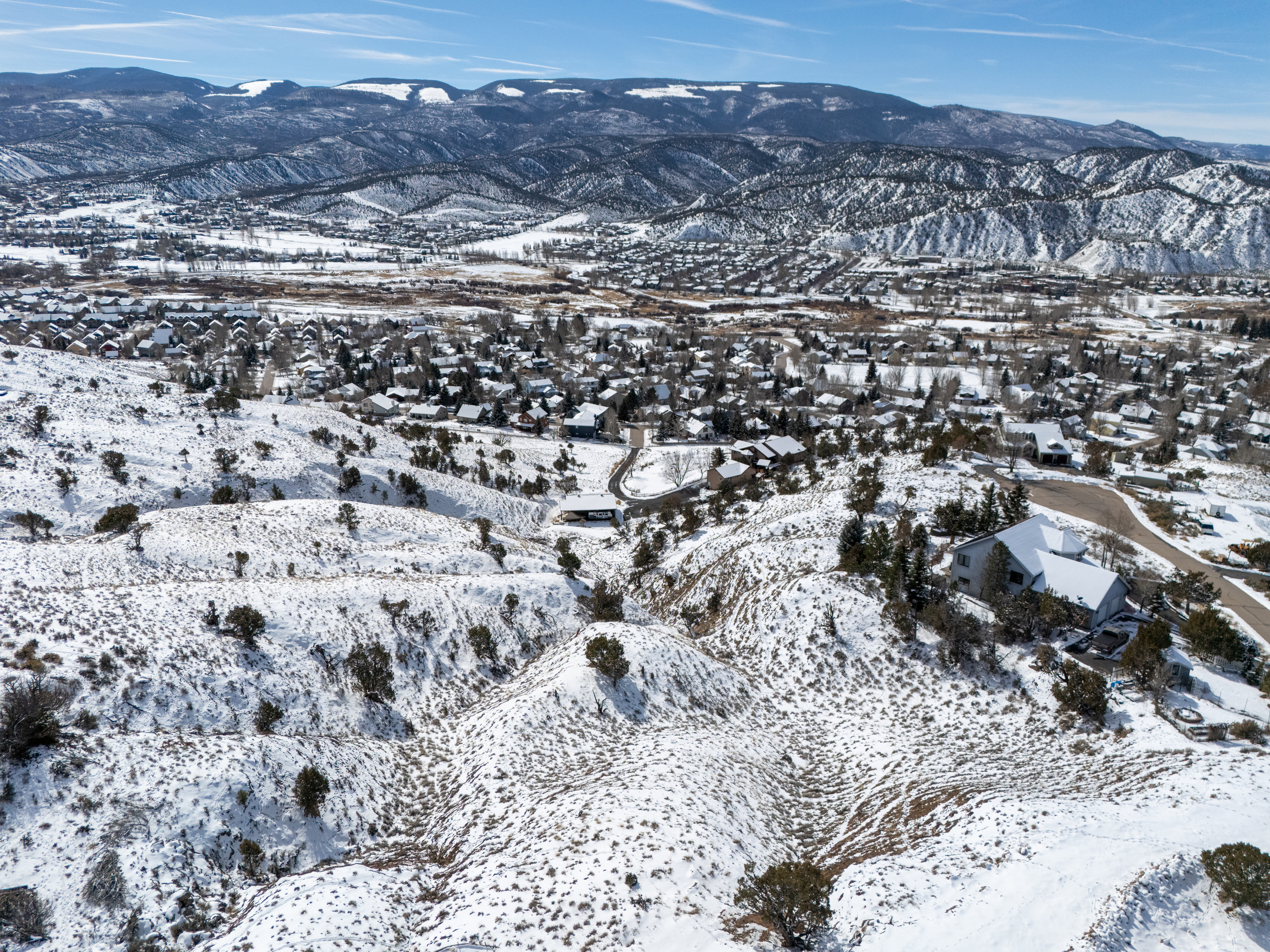 This aerial view showcases a snowy residential area nestled among rolling hills and distant mountains. The houses, seen from above, feature varying rooflines, and the blanket of snow highlights both the natural terrain and the built environment. This image conveys a sense of both seclusion and community within a scenic landscape.
