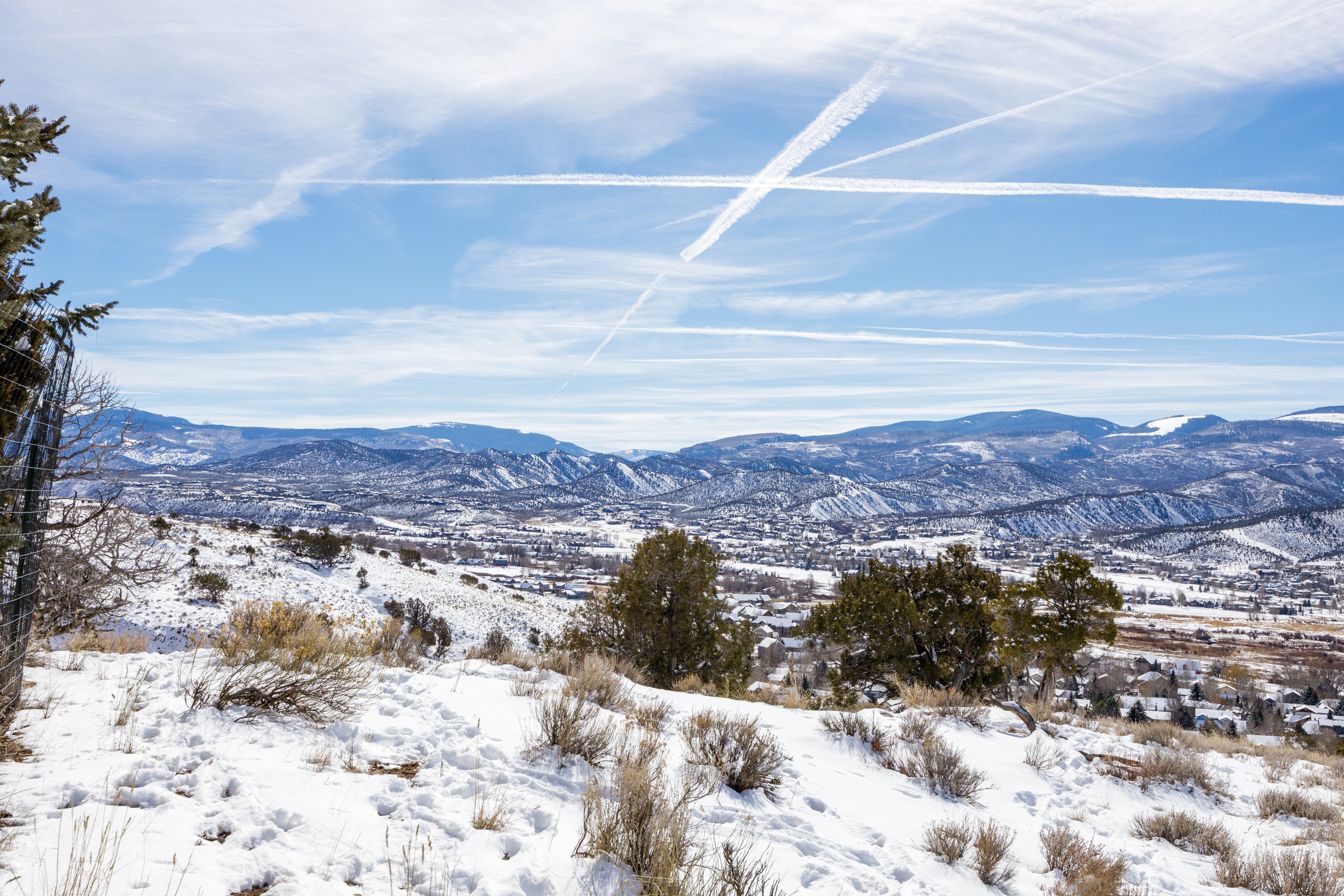 This image offers an aerial view of a snowy landscape encompassing a town nestled between rolling hills and mountains. The foreground features a snow-covered hillside with sparse vegetation, transitioning into a residential area with houses, and further back are the partially snow-covered hillsides and peaks under a blue sky with contrails. This view highlights the property's scenic location and proximity to mountain ranges.