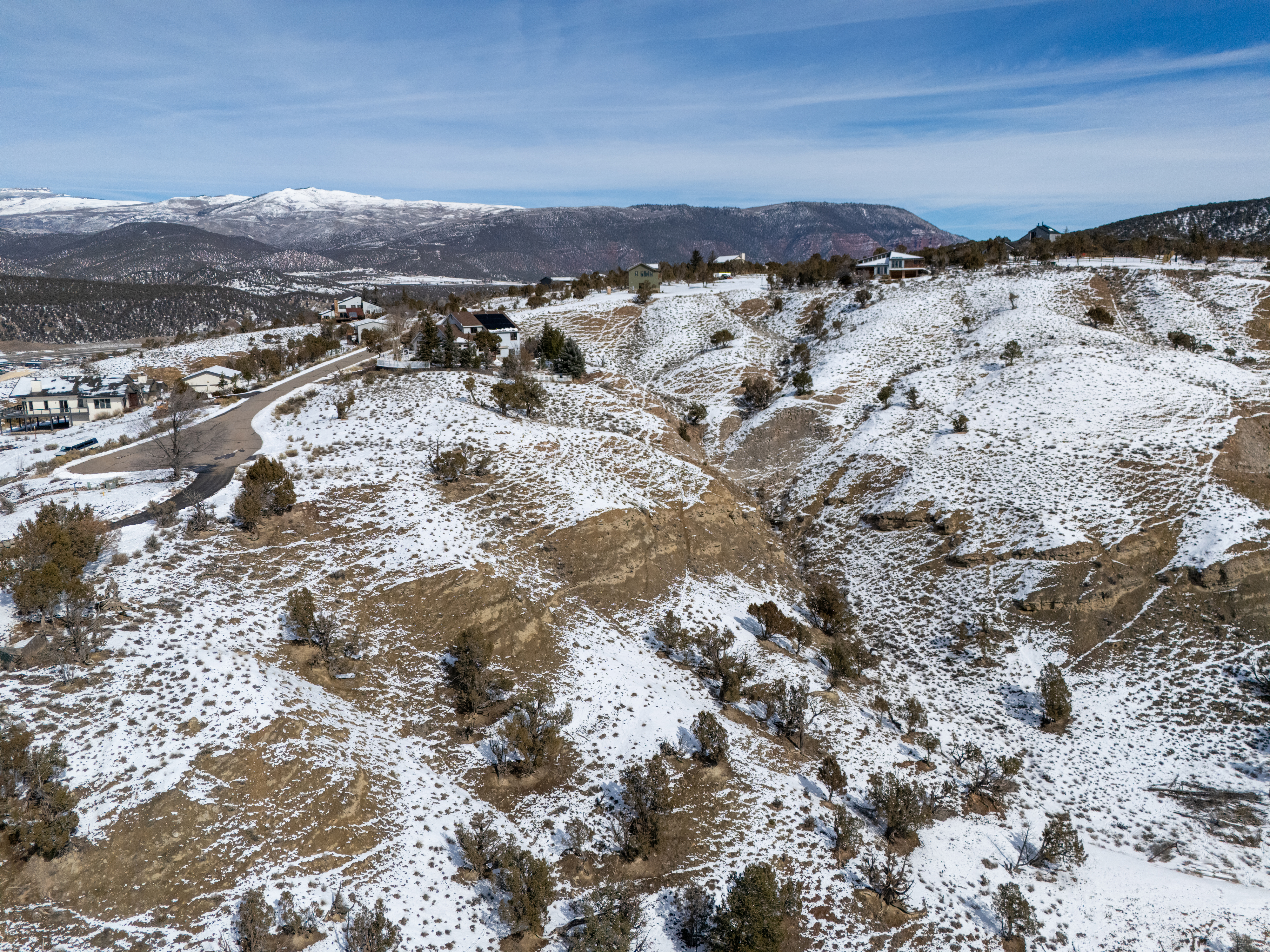 This aerial view showcases a residential area nestled in a snowy, hilly landscape. The homes are situated on the slopes, with some visible roadways connecting them. Mountains can be seen in the background under a partly cloudy sky.