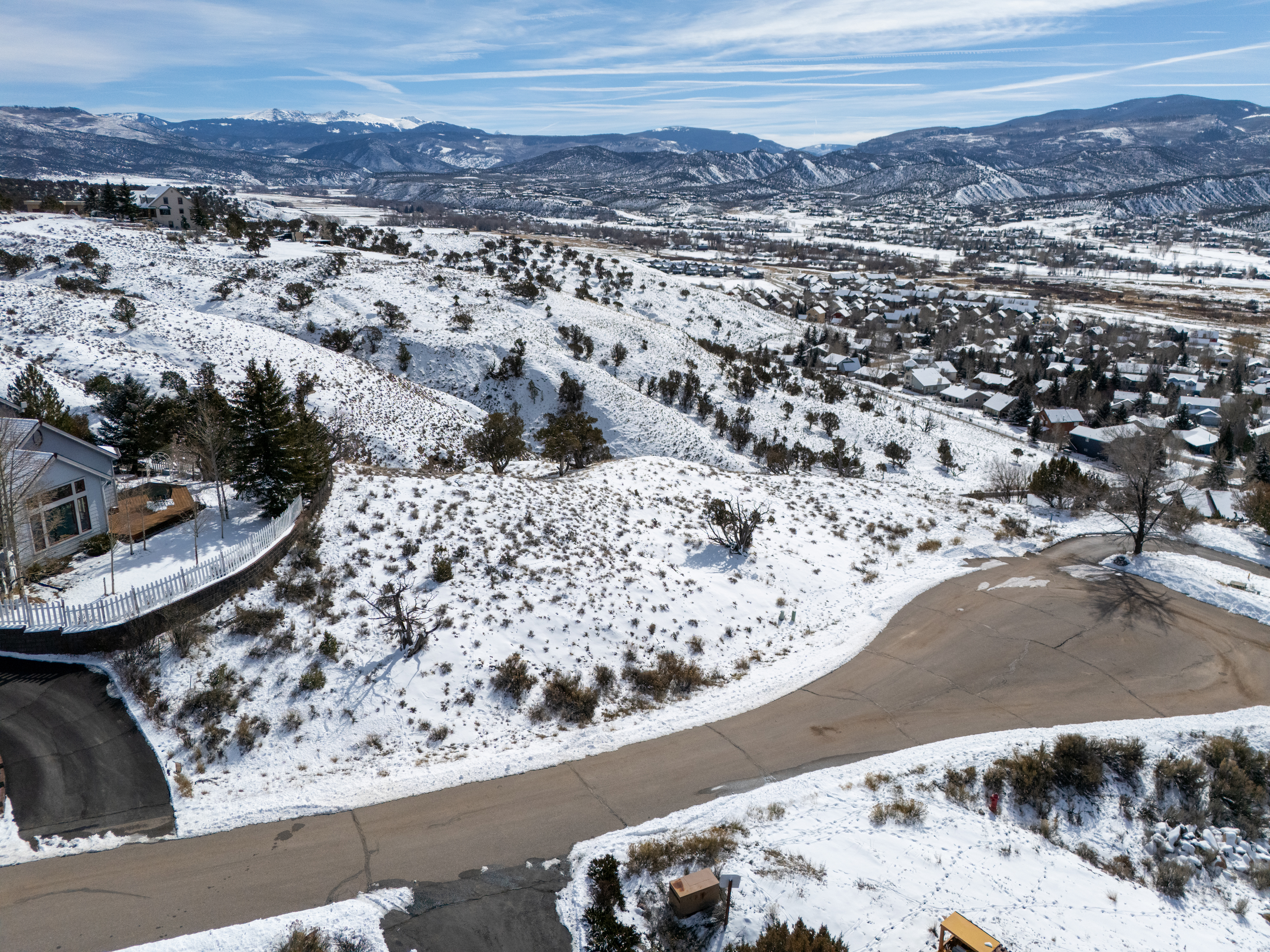 This aerial shot showcases a property nestled in a snow-covered, mountainous landscape. The view features a house with a visible fence and driveway, complemented by a broader perspective of the surrounding residential area and a distant view of the snow-capped mountains. This image emphasizes the property's location and appealing natural setting.