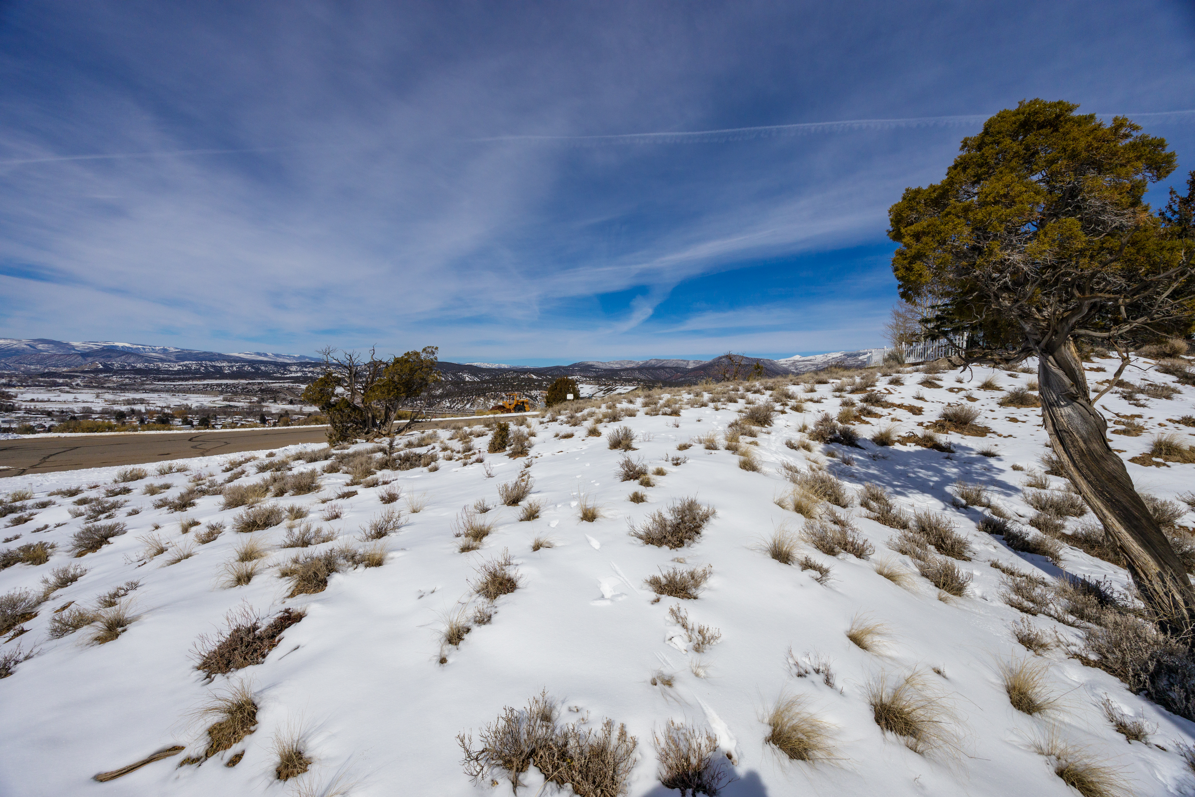 The image shows a snowy hillside landscape, likely part of a property's yard or surrounding area. Patches of snow cover the ground, interspersed with dry vegetation. In the distance, residential areas and snow-capped mountains can be seen, suggesting a scenic location. A gnarled tree stands to the right.