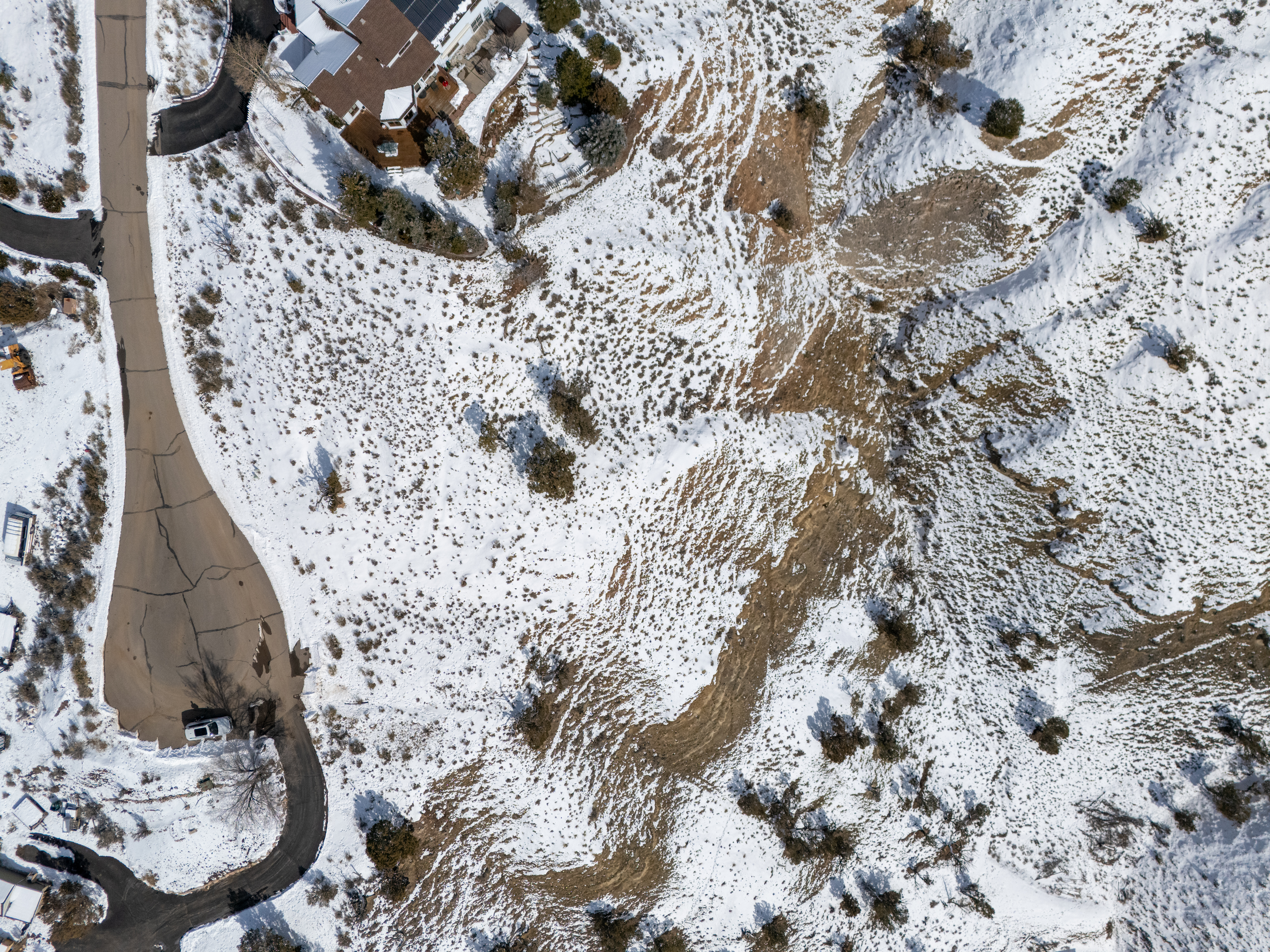 This is an aerial view showcasing a residential property and its surroundings in winter. A paved road leads up to a house with a visible deck and a brown roof partially covered in snow. The surrounding terrain is a mix of snowy patches and exposed ground, with sparse vegetation contributing to the overall wintery landscape.