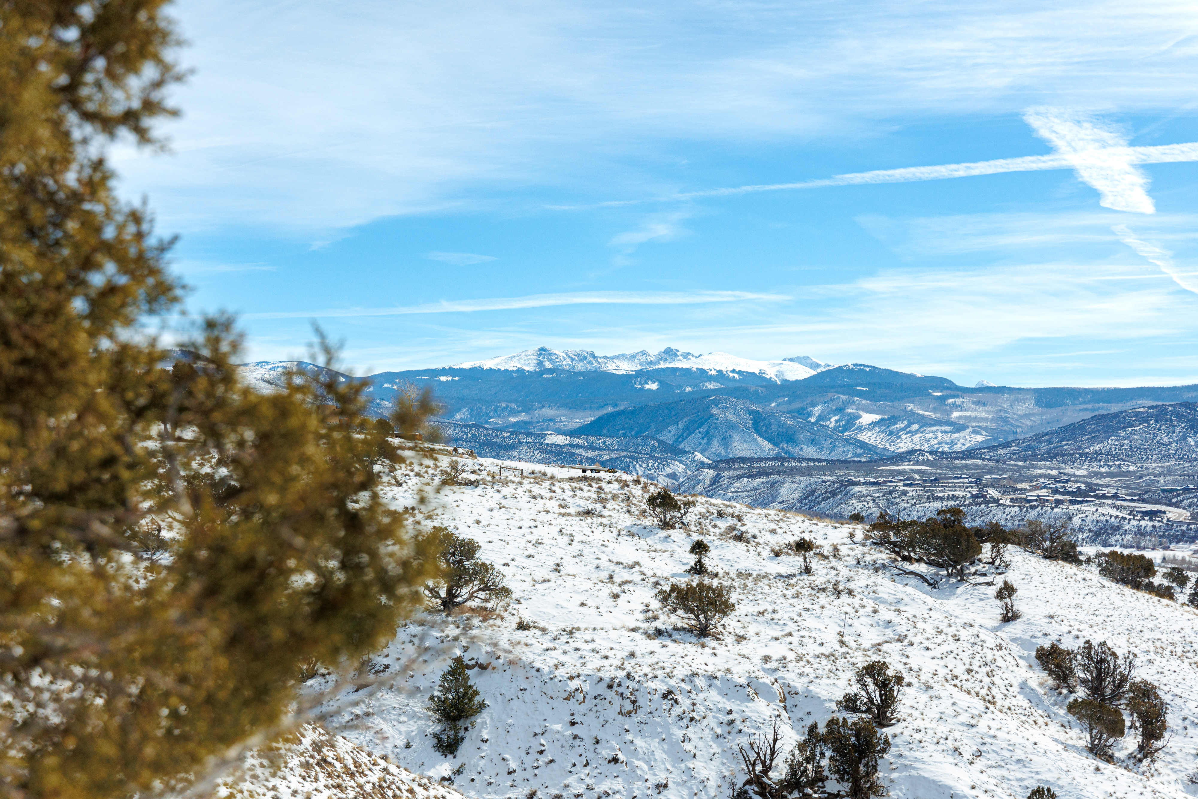 This image showcases a snowy mountain landscape, possibly part of a real estate property. The foreground features a snow-covered hill with scattered trees, while the background reveals majestic, snow-capped mountains under a clear blue sky with contrails. The scene conveys a sense of tranquility and offers stunning natural views, potentially appealing to buyers seeking a secluded mountain retreat.