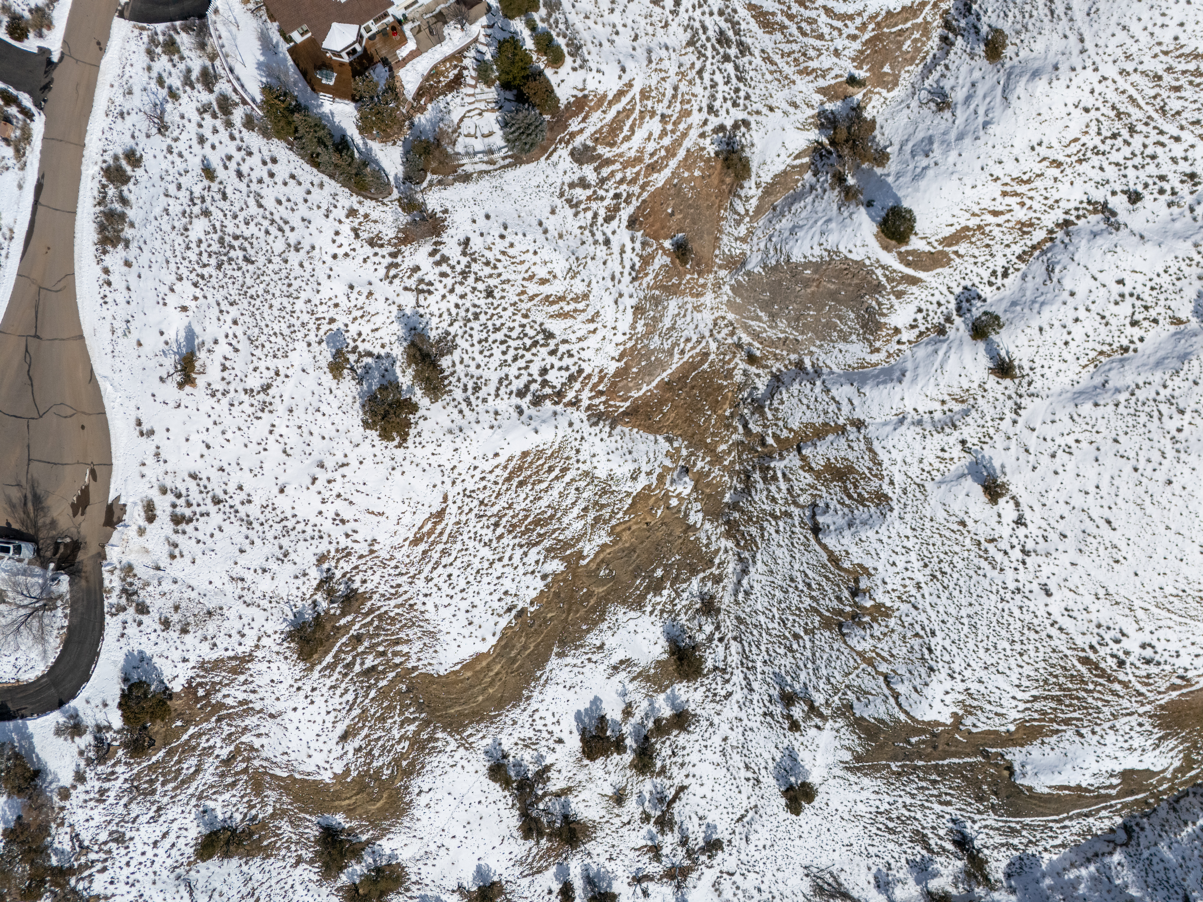This aerial view presents a snow-dusted landscape featuring residential properties and winding roads. The scattered snow cover reveals underlying terrain and sparse vegetation. The properties exhibit varied architectural styles, and the roads suggest a neighborhood setting.