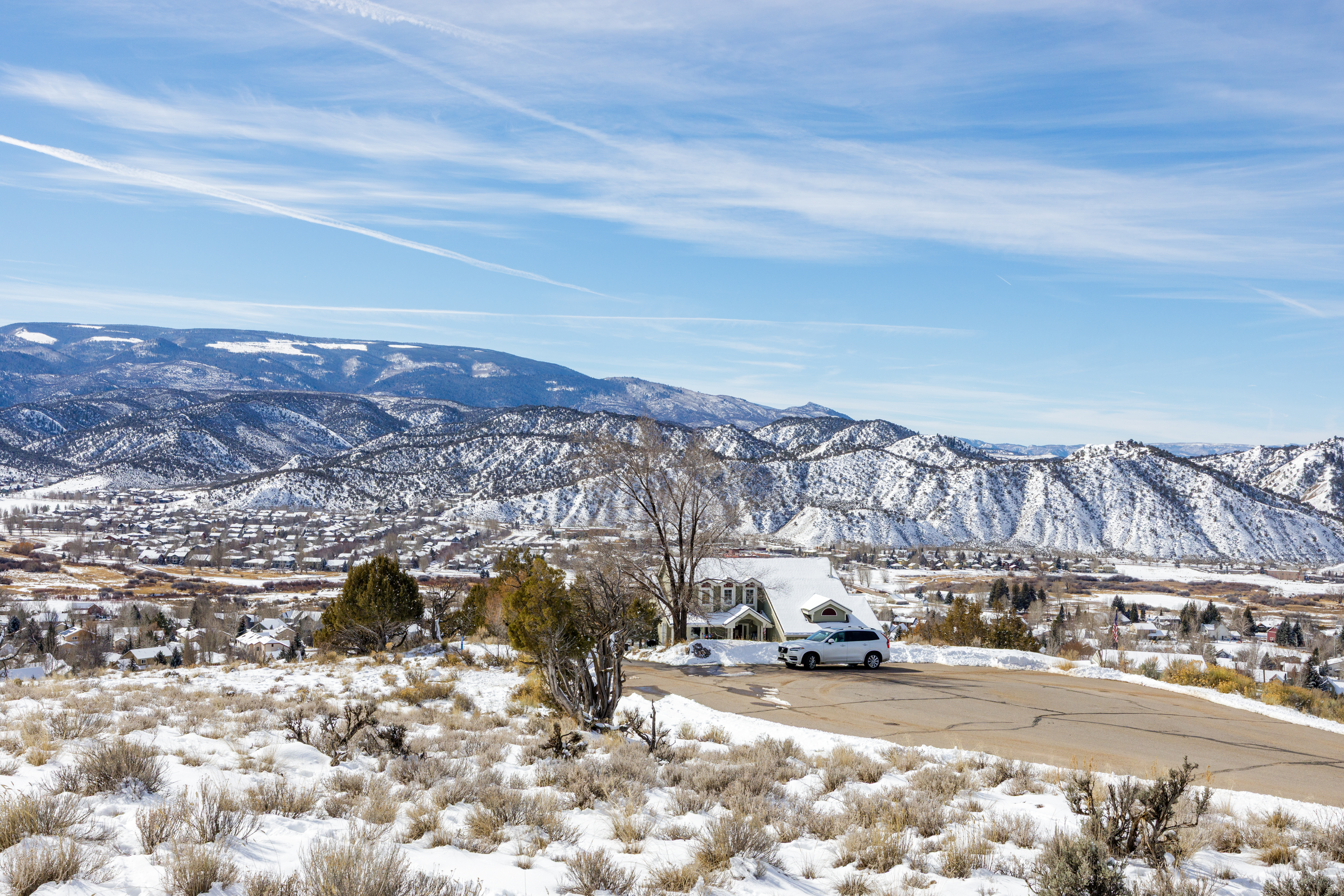 This is an exterior front view of a charming house in a scenic location. Snow covers the ground and rooftops, creating a picturesque winter scene. A white SUV is parked near the house, which overlooks a valley and snow-capped mountains in the distance.